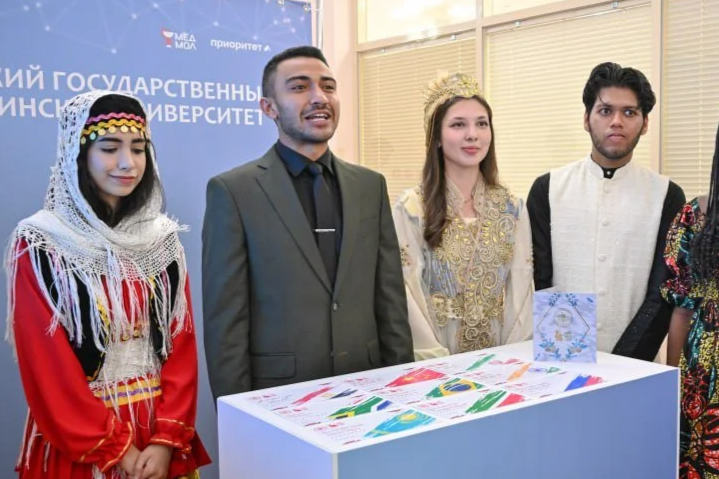 Four people in traditional clothing standing behind a table displaying colorful artwork or crafts at an event, with a blue backdrop and window blinds in the background.