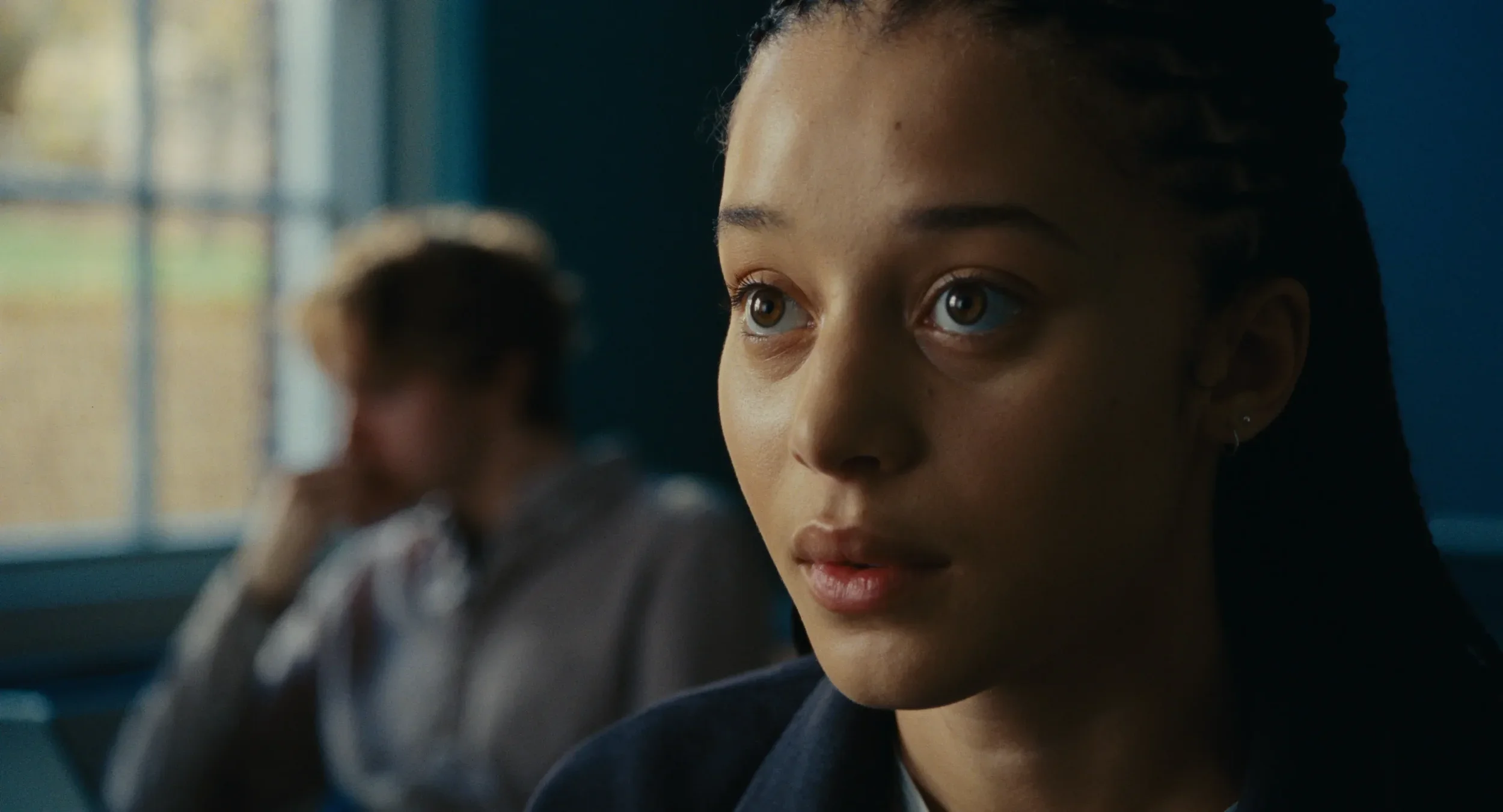 Close-up of a young woman with braided hair and brown eyes, sitting in a classroom, with blurred girl in the background near a window.