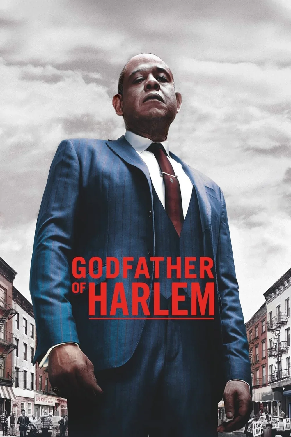 A character wearing a business suit standing with arms at his sides, with a city street in the background and stormy sky overhead, as part of the movie poster for 'Godfather of Harlem.'