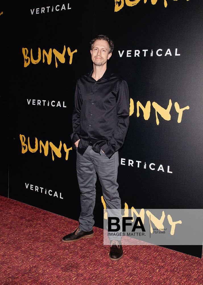 A man standing on a red carpet in front of a black backdrop with the words 'BUNNY' and 'VERTICAL' repeated. He is wearing a black button-up shirt, gray pants, and black shoes.