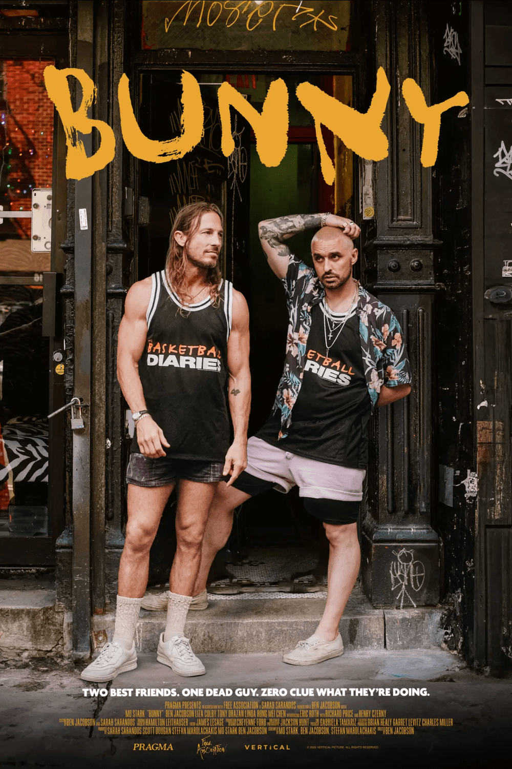 Two men standing outside a black storefront with graffiti, one wearing a basketball jersey and the other a floral shirt with shorts, both looking serious.