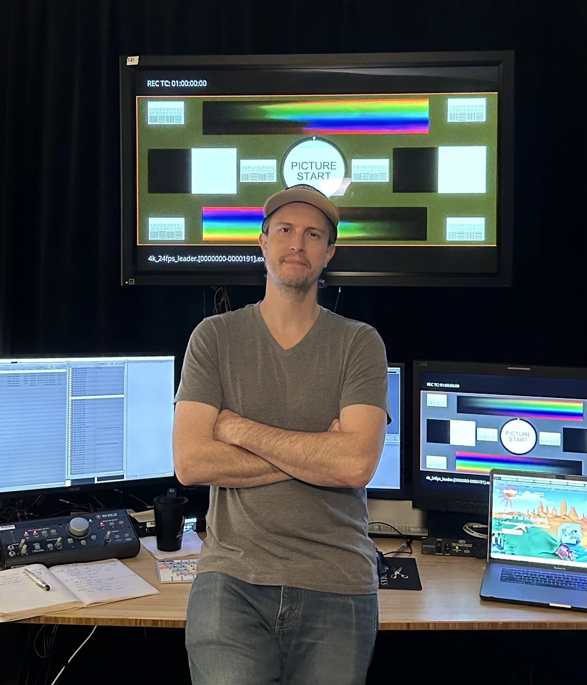 A man with crossed arms standing in front of multiple computer monitors displaying video editing and color calibration screens in a dark room.