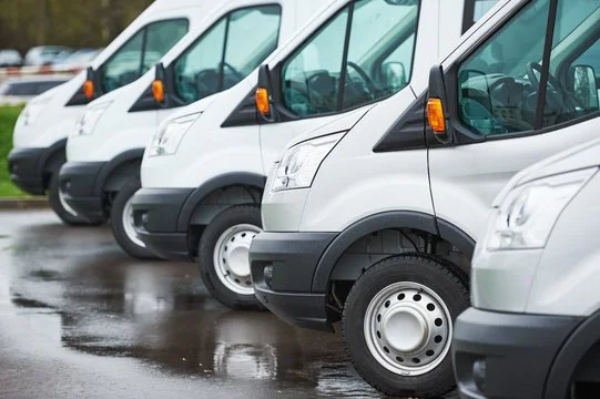 Multiple white delivery vans parked in a row on a wet surface.