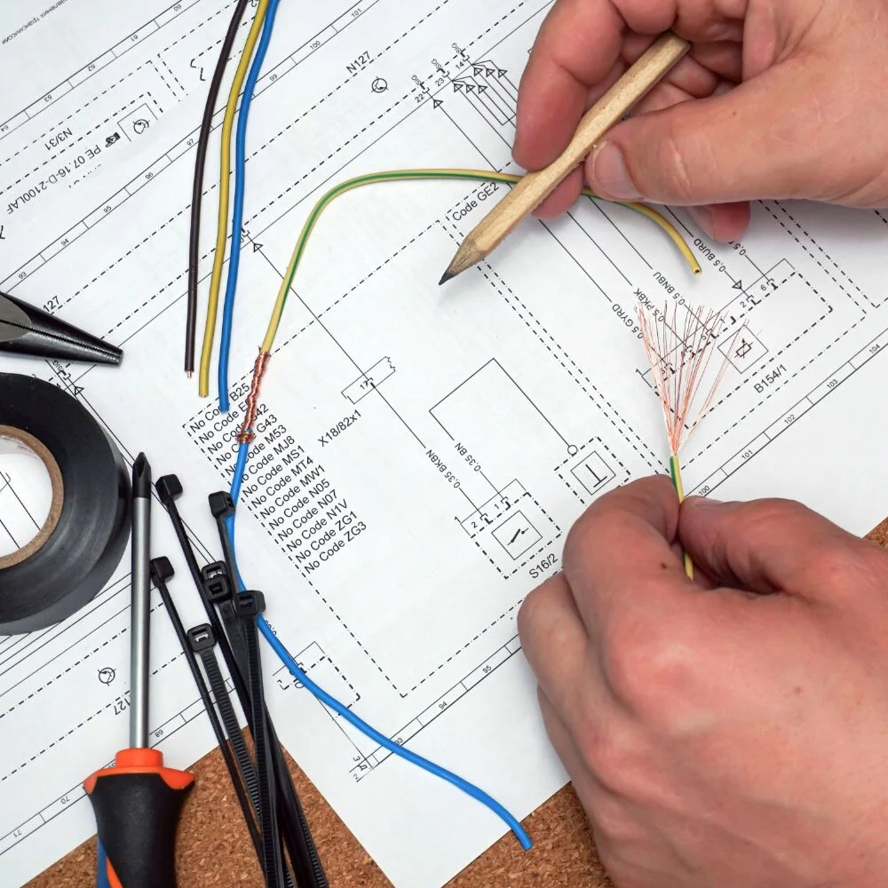 Person wiring electrical cables on a technical blueprint or wiring diagram, with tools and electrical tape nearby.