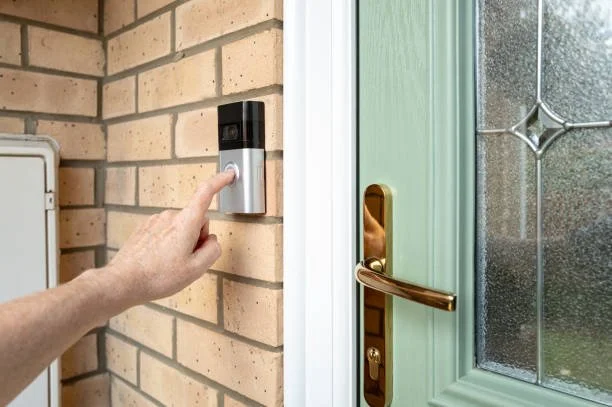 A person pressing a doorbell button on a brick wall outside a house with a green door and gold handle.