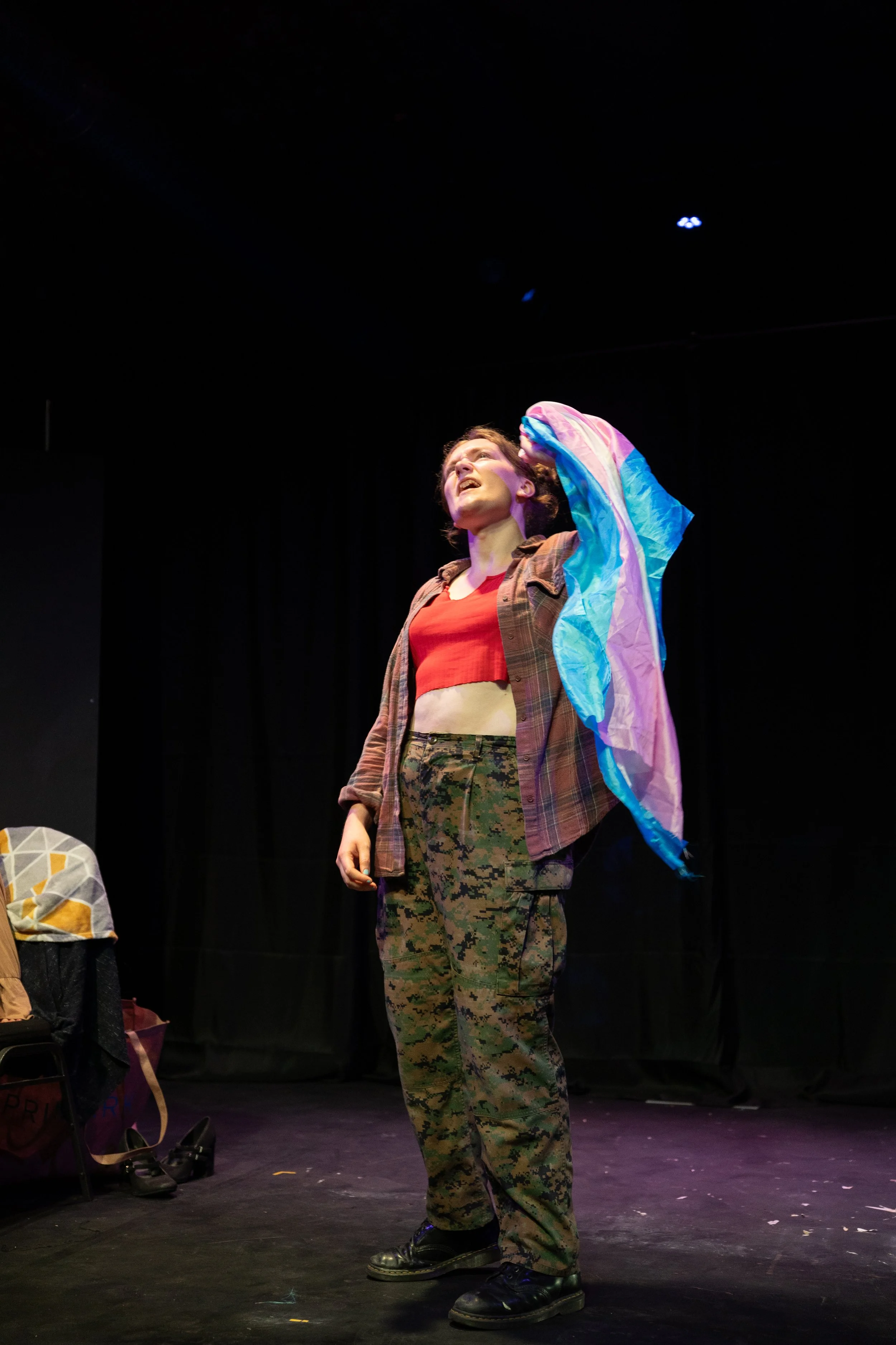 A woman in camouflage pants, a red crop top, and a plaid shirt holding a colorful flag, standing on a stage with a black curtain background.