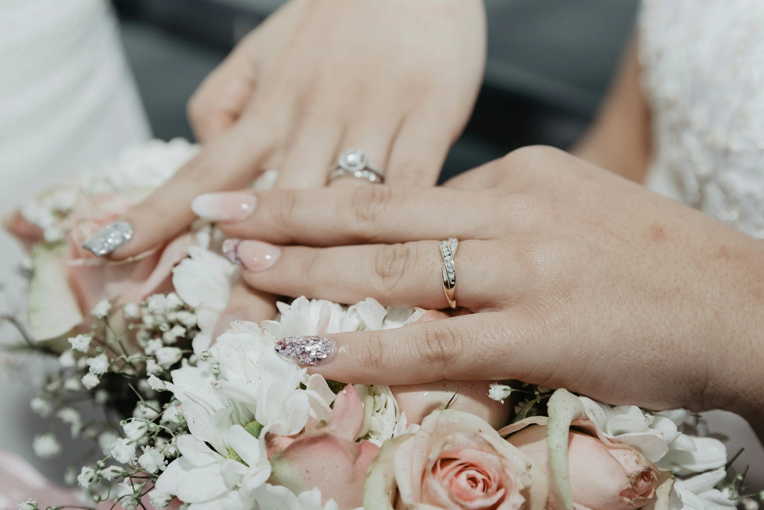 Close-up of two hands with wedding rings on fingers, surrounded by a bouquet of white and blush pink flowers.