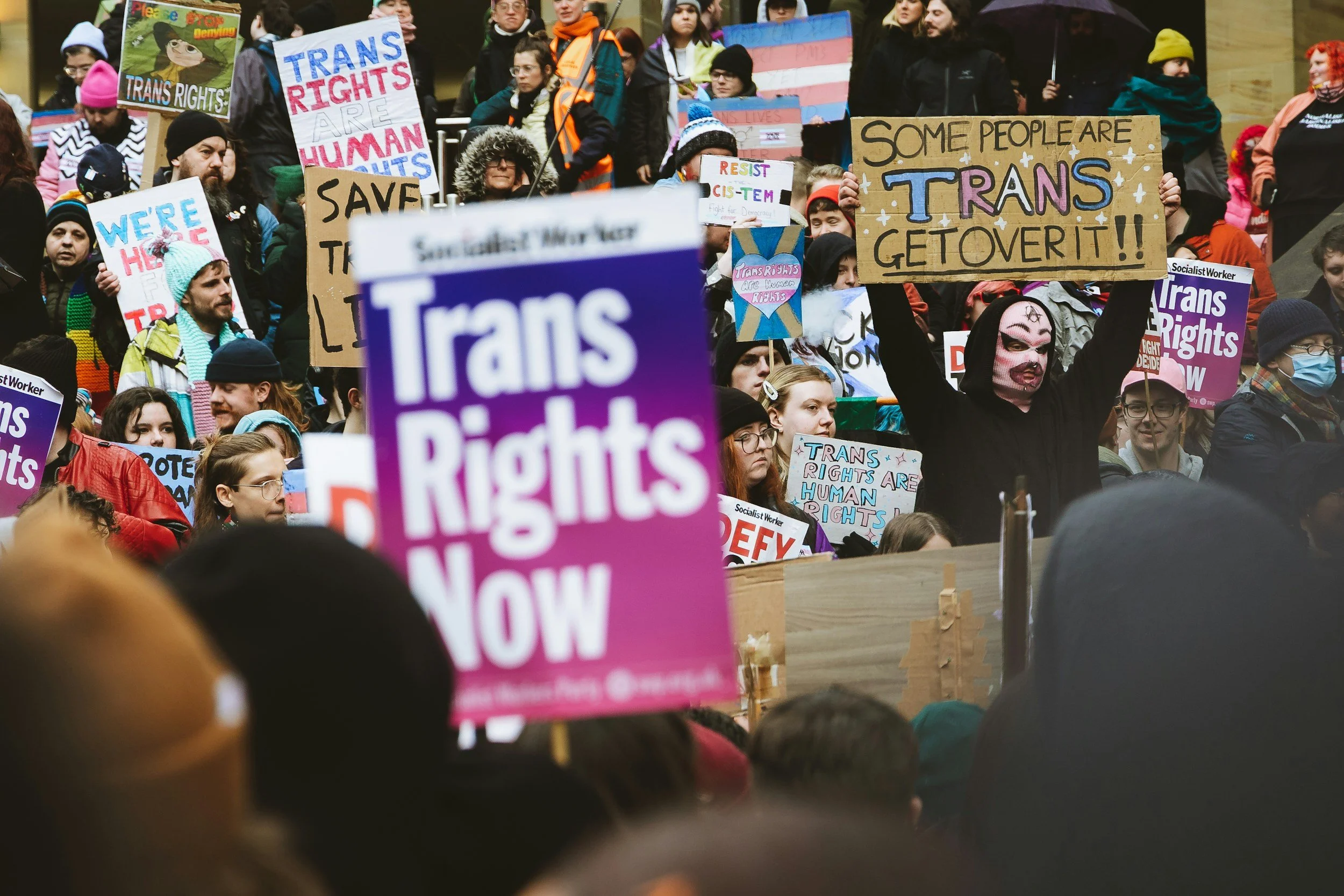 Crowd of people at a protest holding signs advocating for trans rights and LGBTQ+ equality. Some signs read 'We're here,' 'Save trans,' 'Some people are trans, get over it,' and 'Trans rights are human rights.' The protesters are dressed warmly, indicating cold weather.