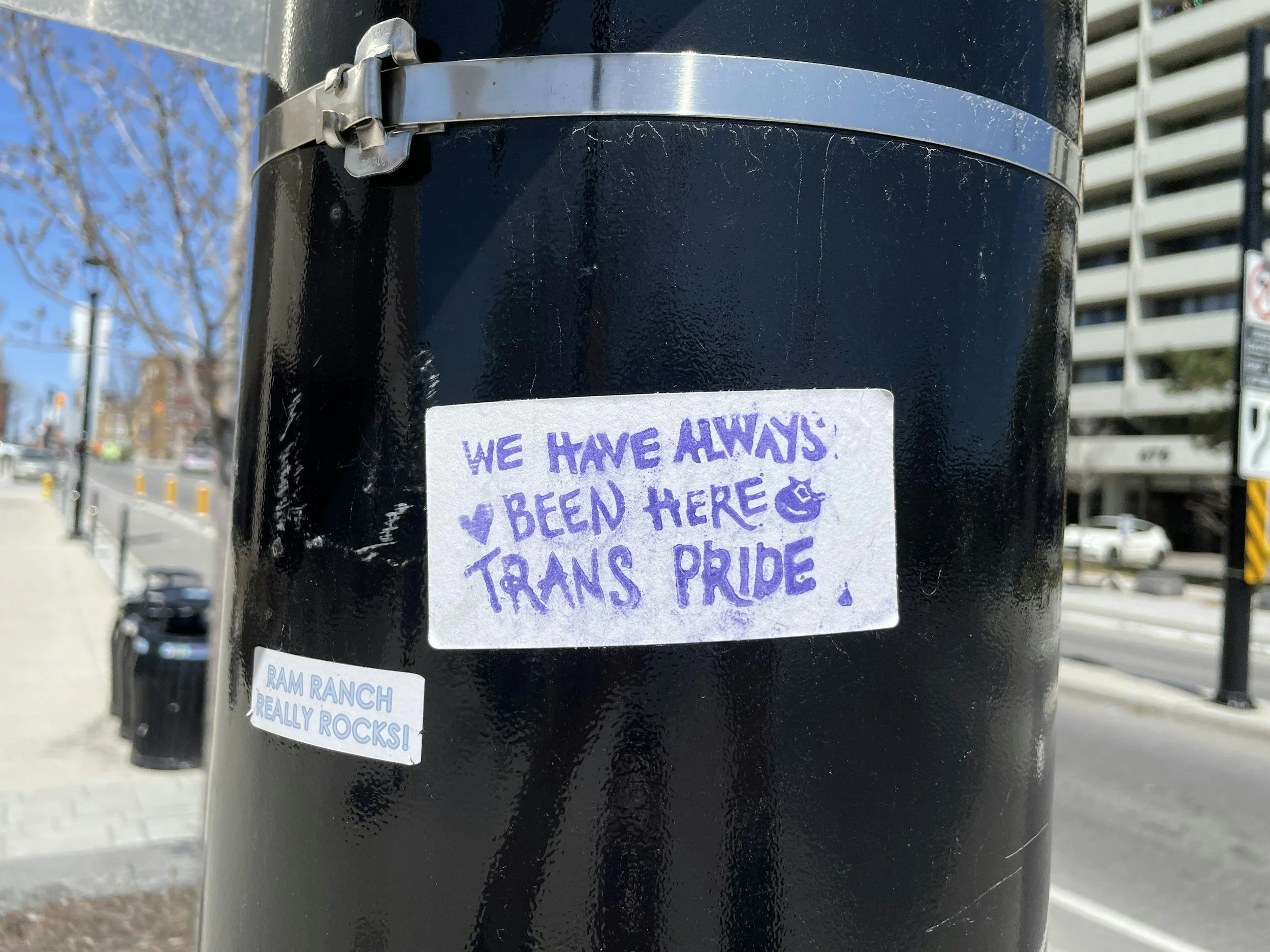 Close-up of a black pole or post with two stickers and a handwritten note, with a background of buildings and trees on a sunny day.