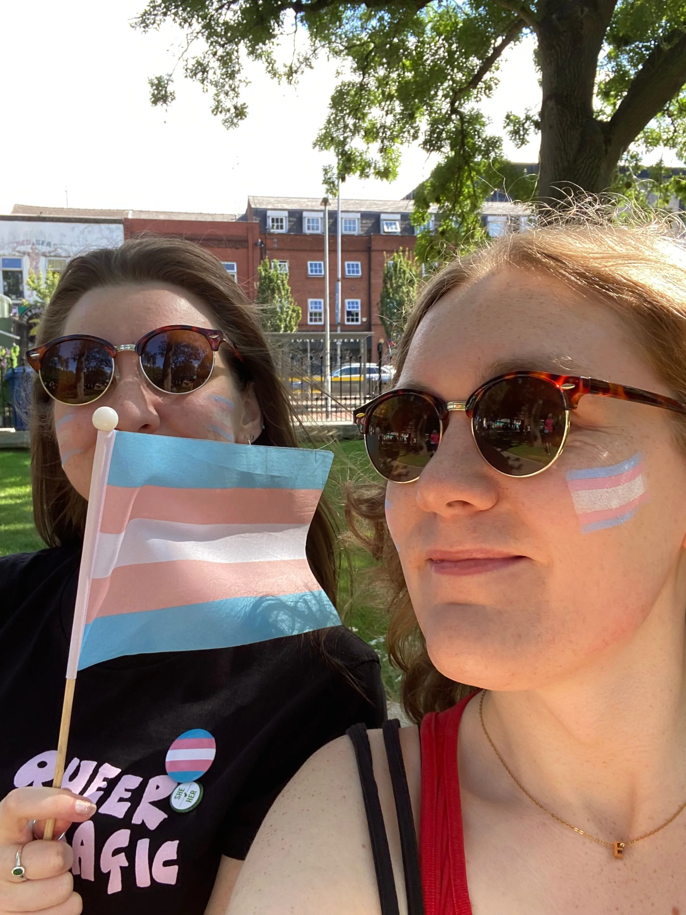 Two women wearing sunglasses and face masks holding queer pride flags and pins, standing outdoors near a large tree with a building in the background during daytime.