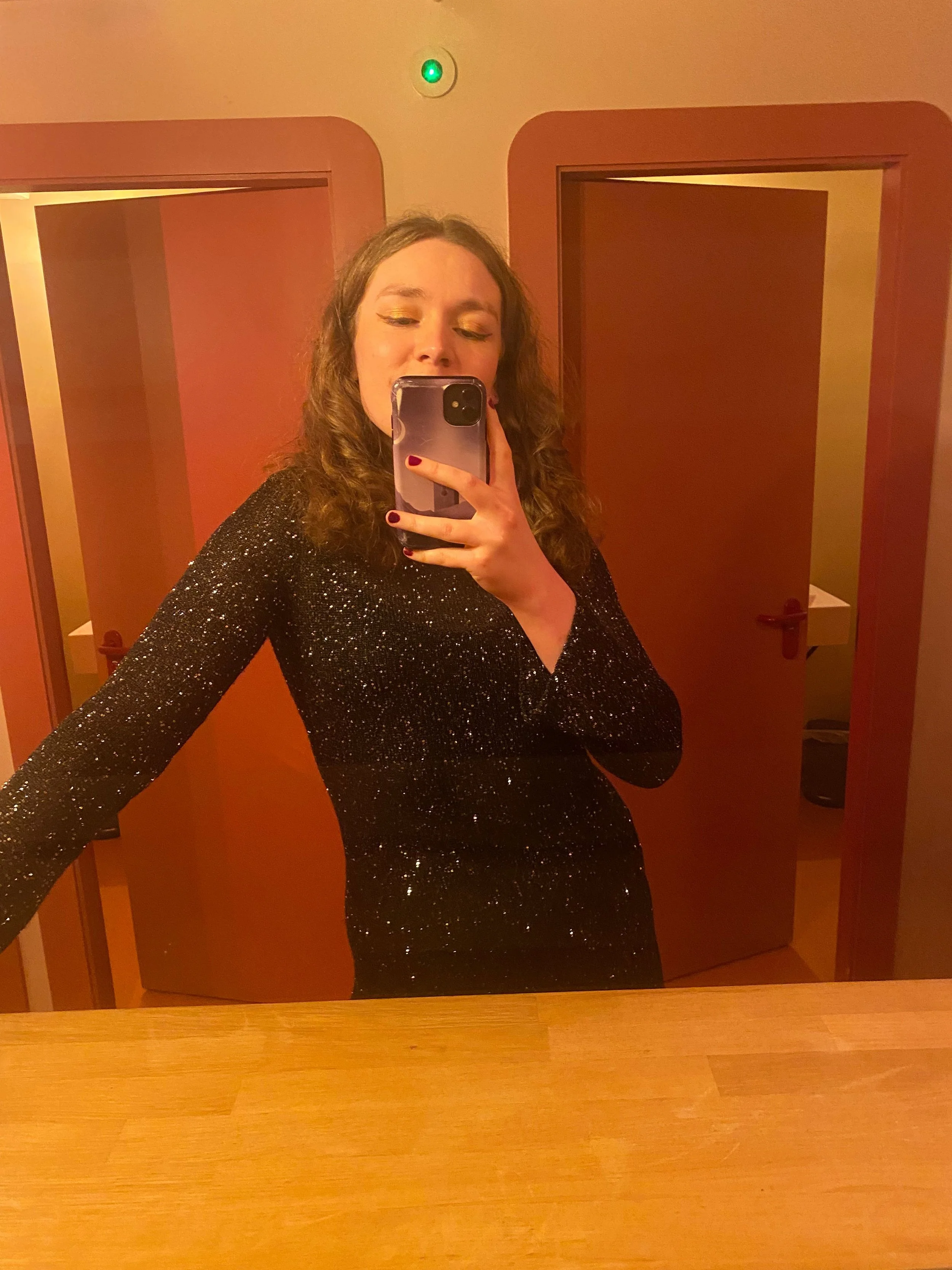 A woman with brown, curly hair taking a mirror selfie in a room with wooden doors. She is wearing a black, glittery, long-sleeve dress and has red nail polish.