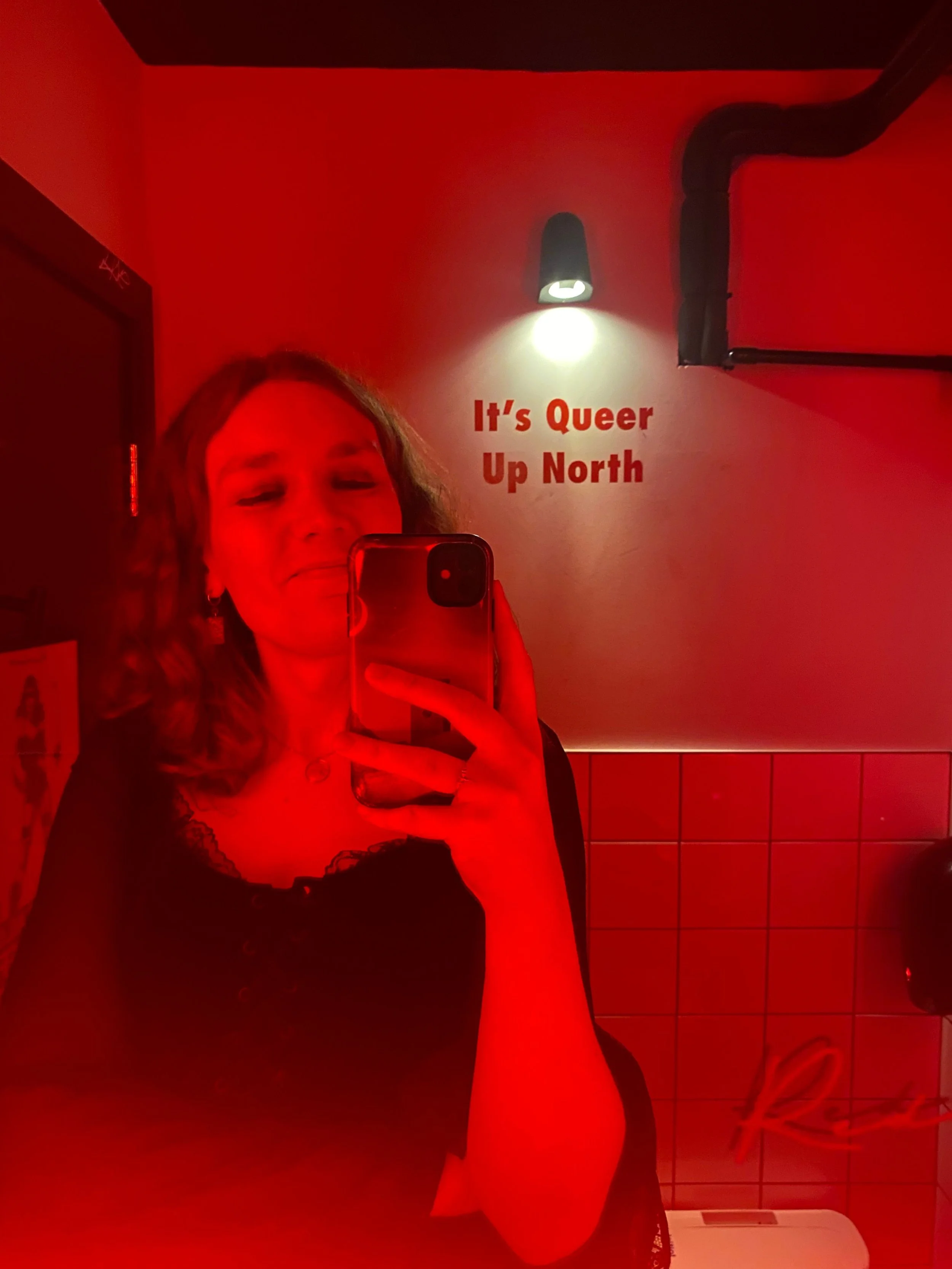 A person taking a selfie in a mirror at a restroom with red lighting. There is a wall with a sign reading 'It's Queer Up North' and some black pipes are visible.