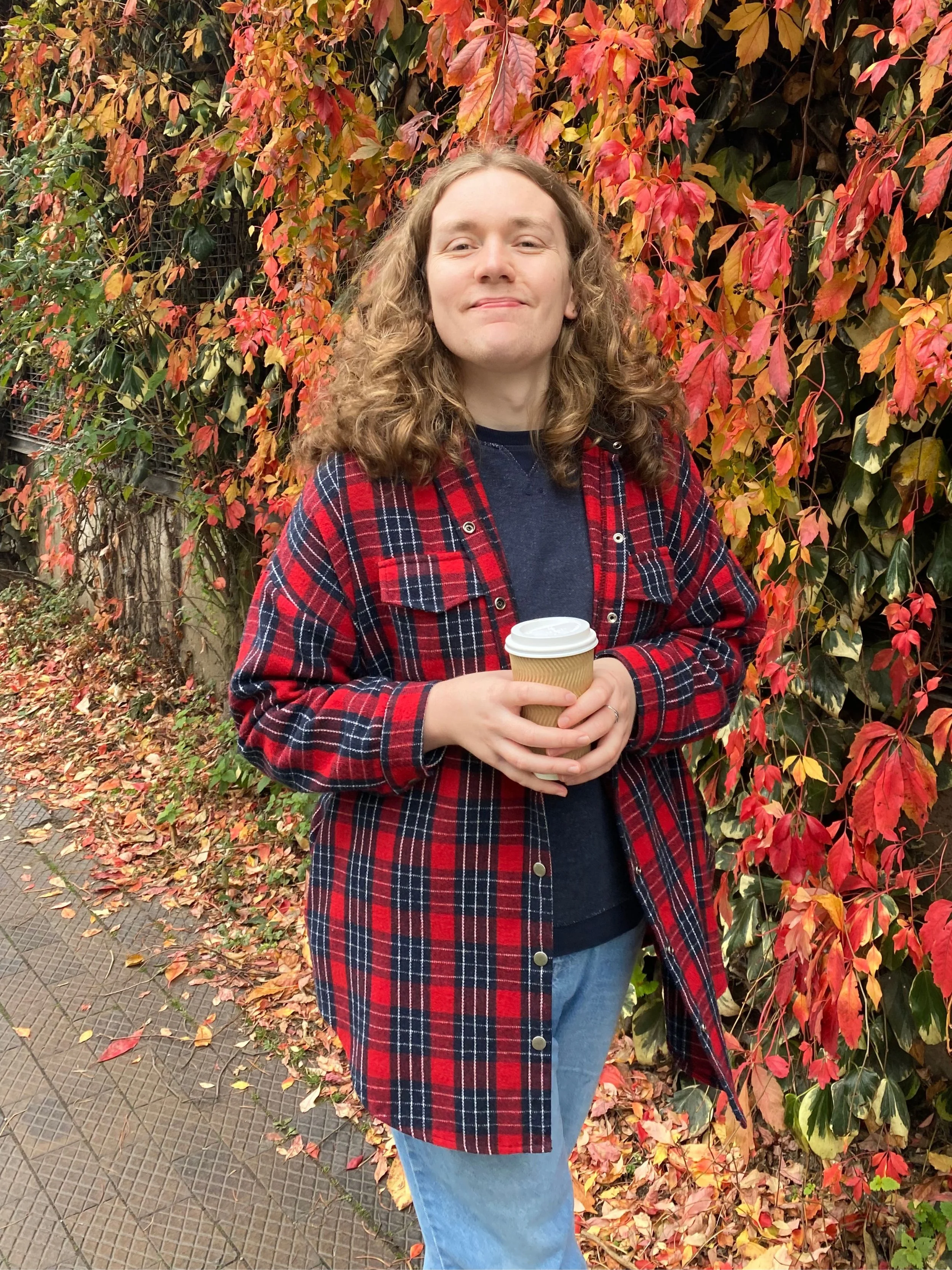 A woman with curly hair stands outdoors in front of a wall of colorful autumn leaves, holding a disposable coffee cup, wearing a black shirt and red plaid jacket.