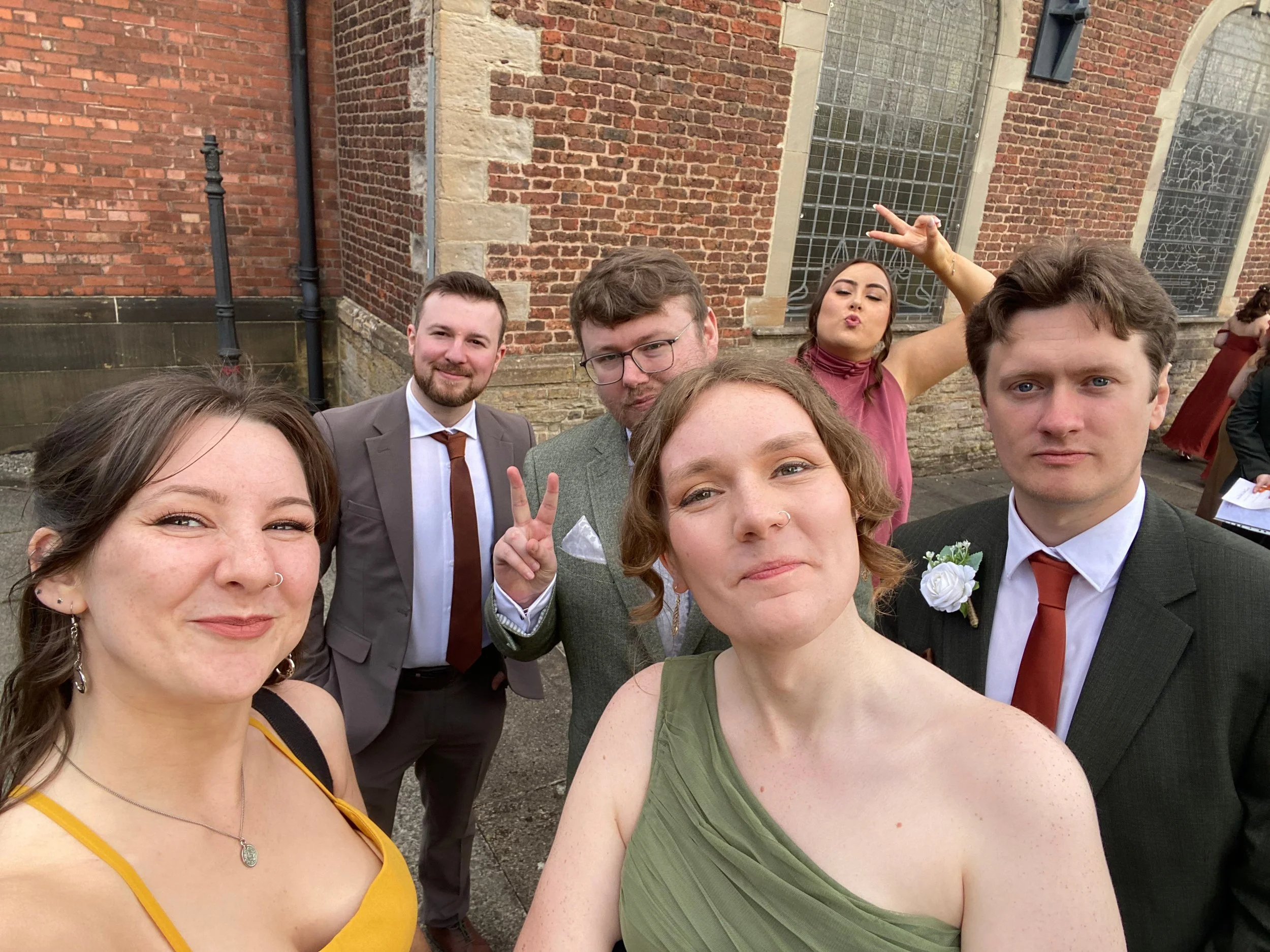A group of five young adults taking a selfie outside a brick building during daytime. The people are dressed in semi-formal attire, with some smiling and one making a peace sign. The background features brick walls and windows with decorative metalwork.