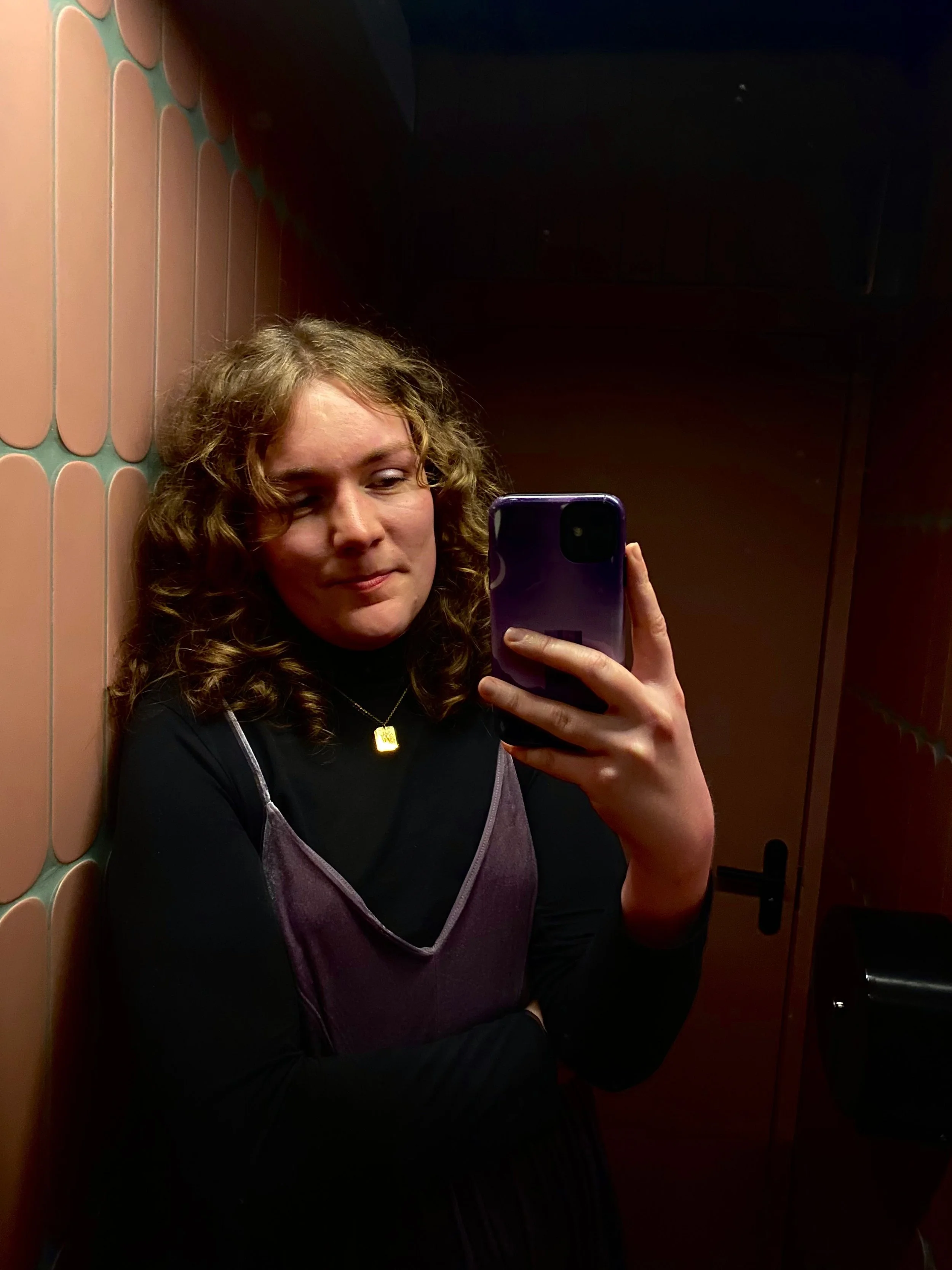 A young woman with curly hair taking a selfie in what appears to be an elevator mirror, wearing a black long-sleeve shirt and a purple sleeveless top with a gold necklace, standing against a background with peach-colored tiles and dark brown walls.