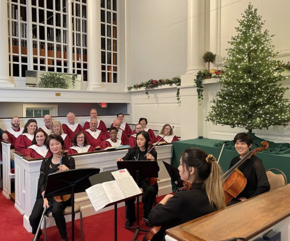 Choir and string musicians performing inside a church decorated for Christmas with a lit Christmas tree and holiday garlands.