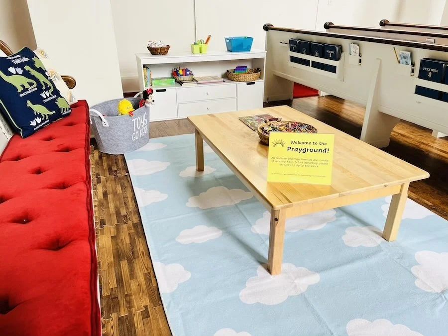 Indoor playground area with a red couch on the left, a light blue rug with white clouds on the floor, a wooden table in the center, a white storage unit with toys and books in the background, and a yellow sign on the table that says "Welcome to the Playground!"