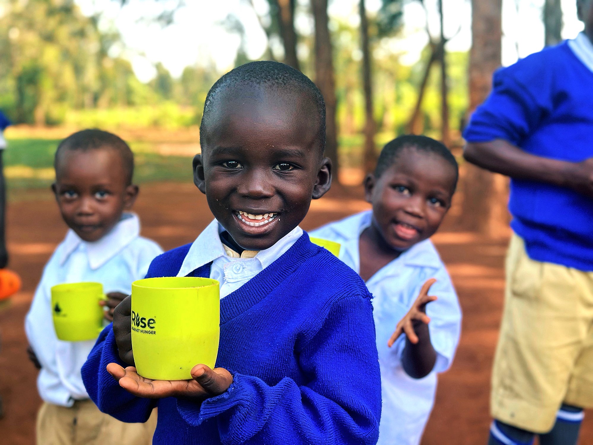 Young African boy in a blue school uniform smiling and holding a yellow charity cup with the words "Rise Against Hunger" written on it, standing outdoors with other children in the background.