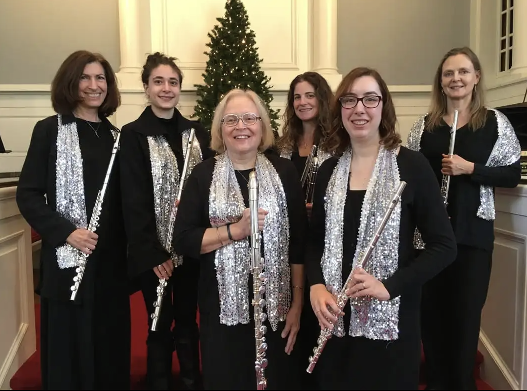 Group of six women holding flutes in front of a decorated Christmas tree indoors.