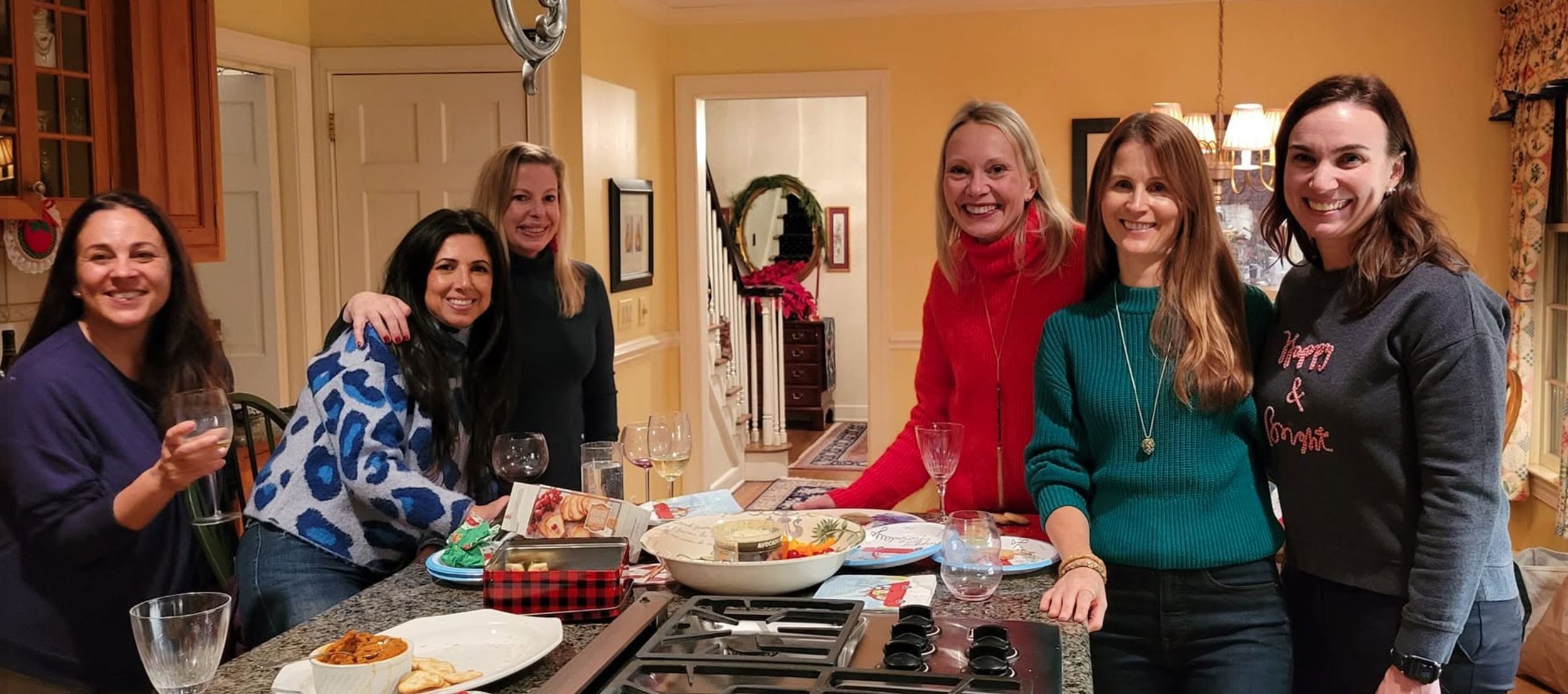 Group of women smiling and posing in a kitchen during a holiday celebration, with food and drinks on the table.