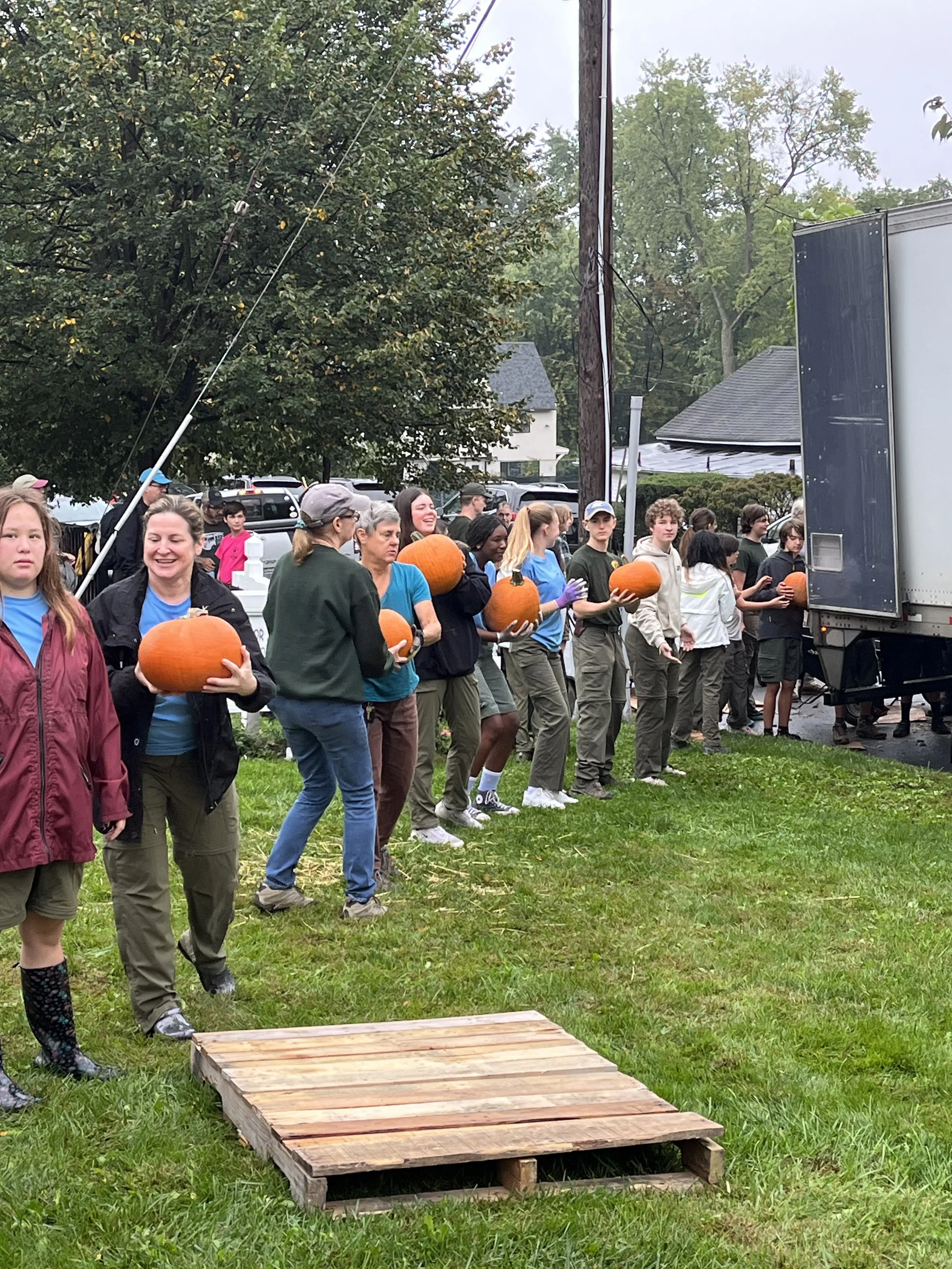 People standing in a line outdoors holding pumpkins, with a wooden platform on the grass in front of them, and a truck nearby.
