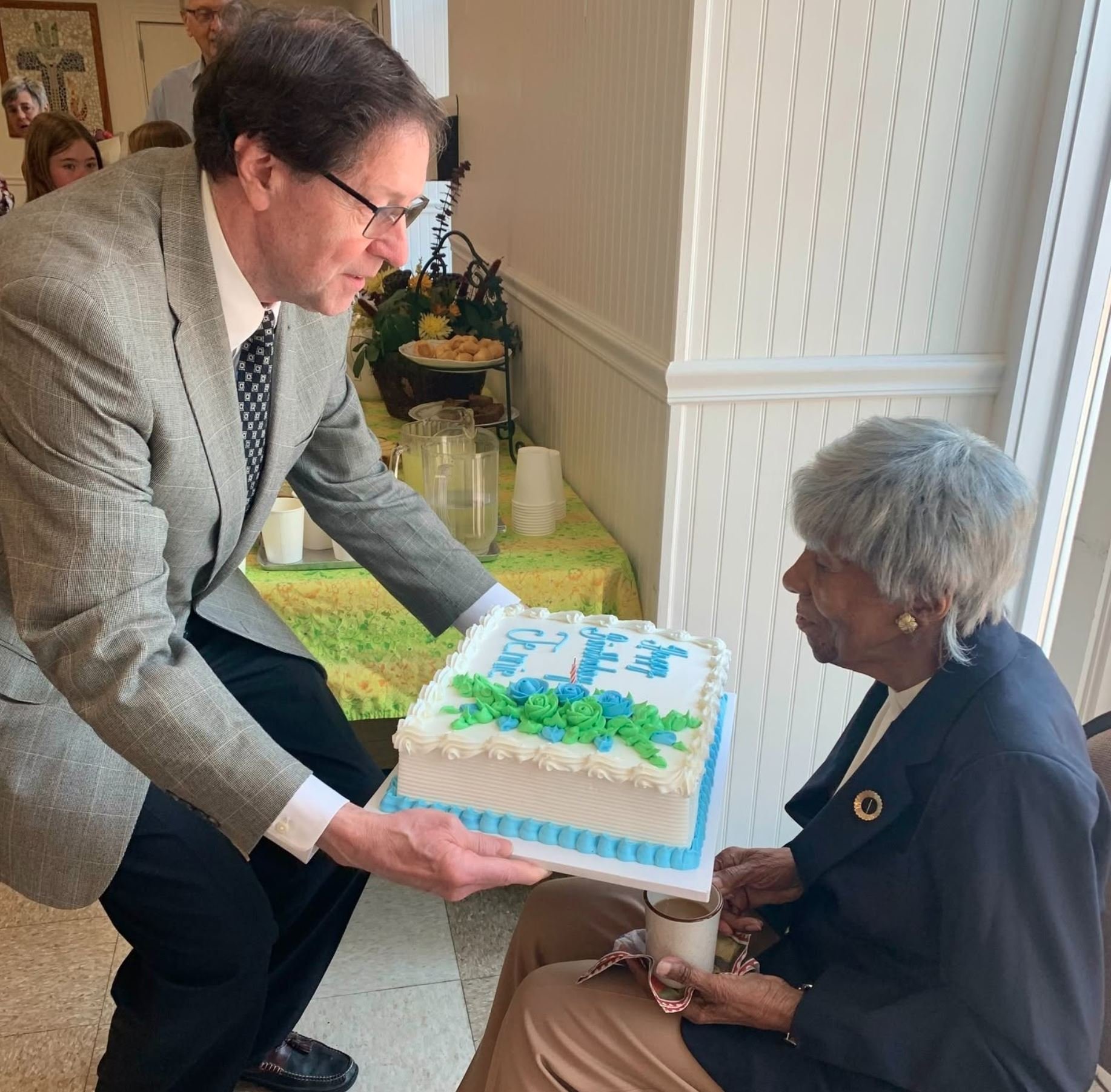 A man in a gray suit presenting a decorated birthday cake to an older woman who is celebrating her 100th birthday