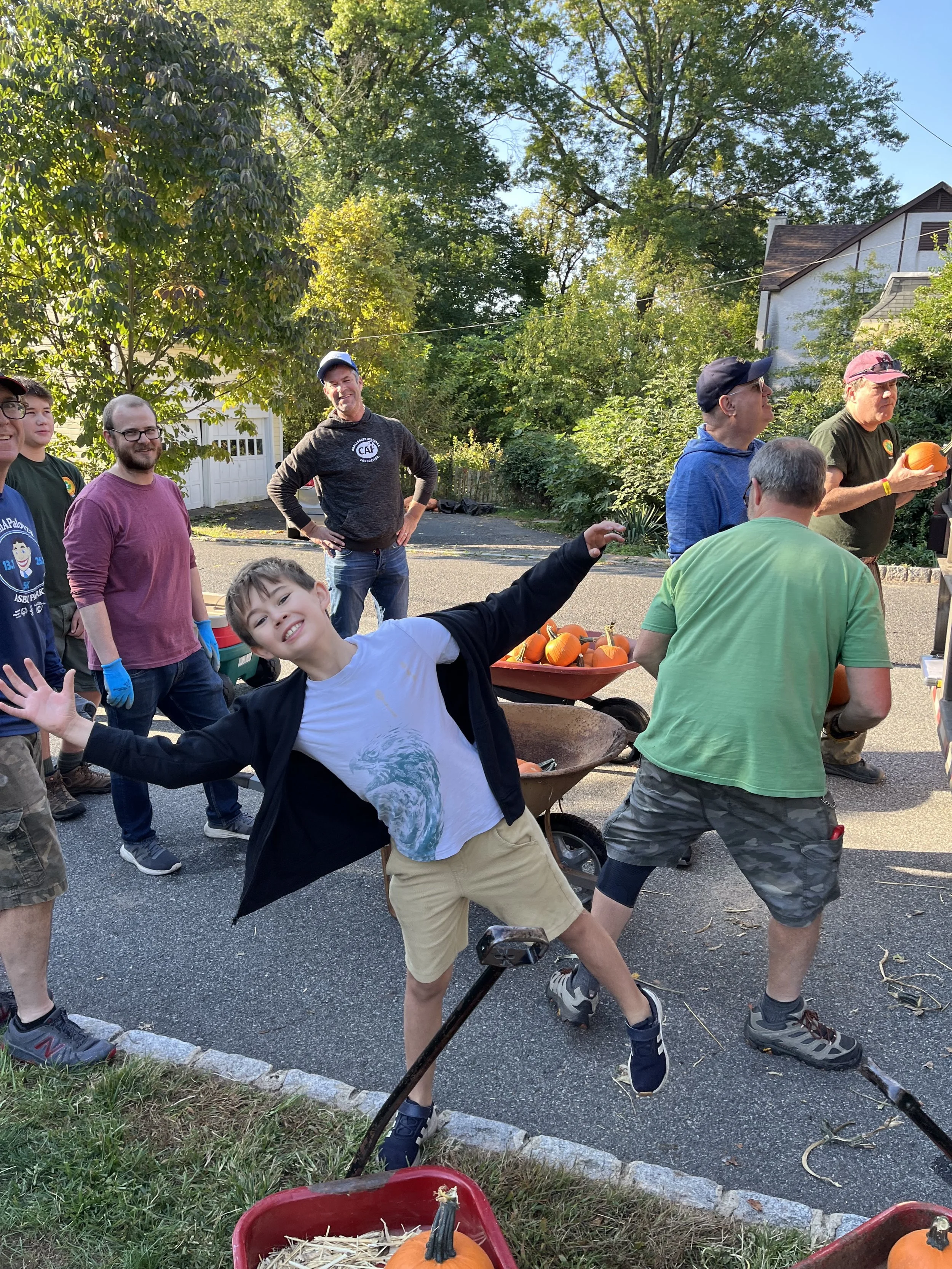 A group of people gathered outdoors during daytime, celebrating Halloween with pumpkins. A young boy in the foreground strikes a playful pose with a big smile, while adults in the background stand and chat, some holding or near pumpkins. The setting includes trees, a house, and a wheelbarrow filled with pumpkins.