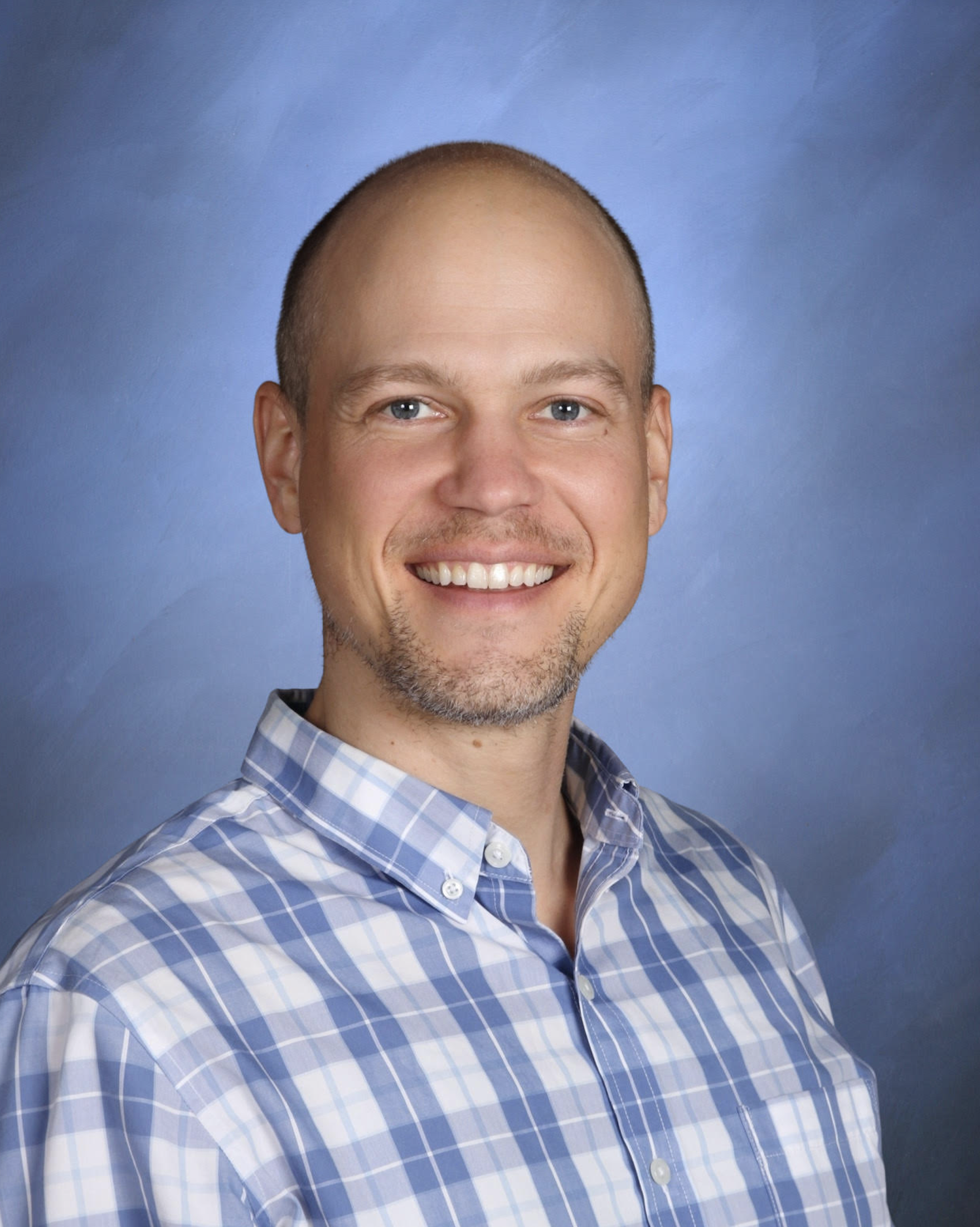 Close-up portrait of a smiling man with short hair and a beard, wearing a blue and white checkered shirt, against a blue background.