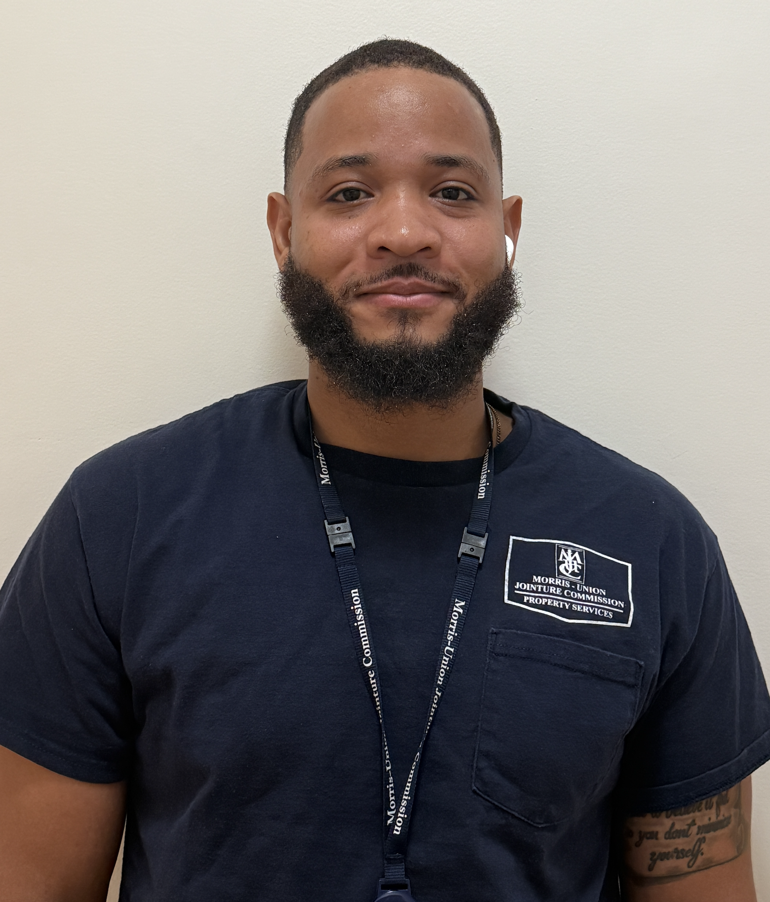 A man with short hair and a beard, wearing a navy blue T-shirt with a badge that reads 'Morris-Union Jointure Commission Property Services,' standing against a plain white wall.