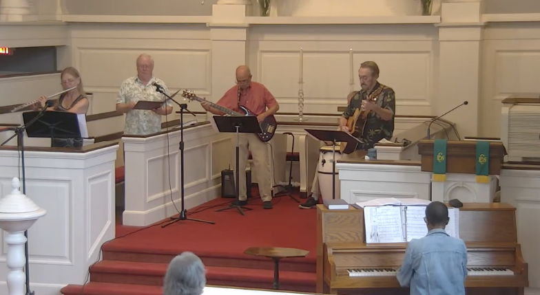 A group of four musicians performing in a church with an audience. The band includes a woman playing the flute, a man singing and playing an acoustic guitar, a man playing an electric guitar, and another man playing the keyboard. A person on piano is in the foreground, facing the band, with sheet music open in front of him.