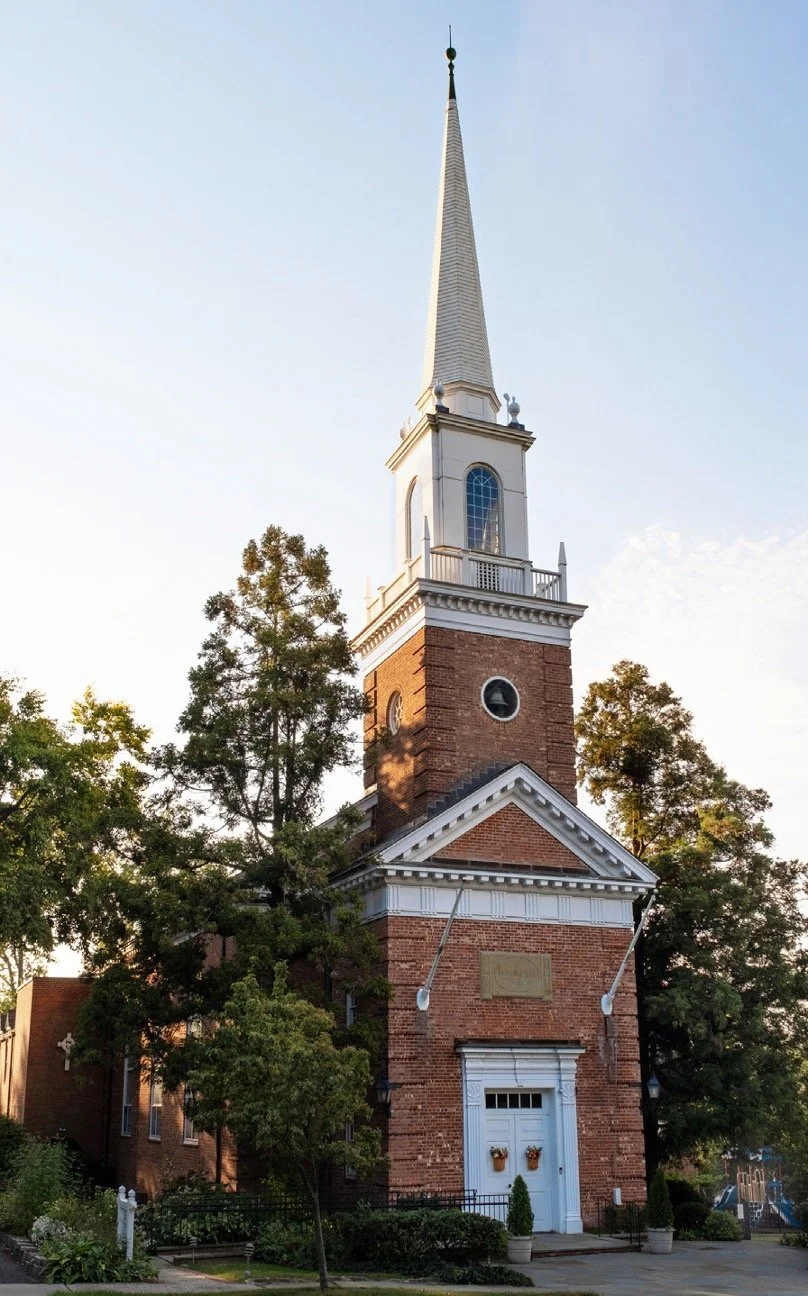 A historic red brick church with a tall white steeple, surrounded by trees and a small garden, under a clear sky.