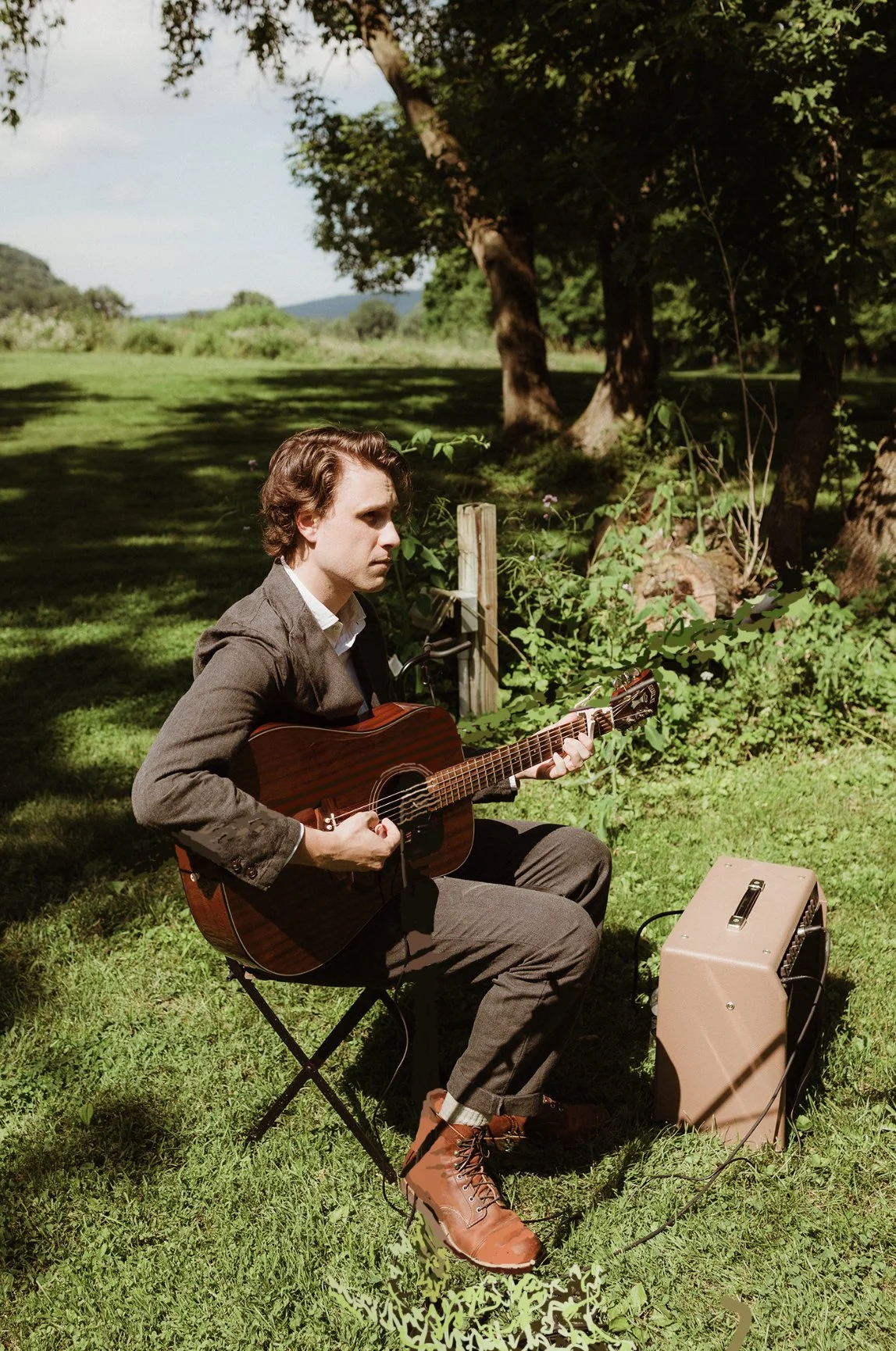 A young man with wavy brown hair playing an acoustic guitar outdoors on a grassy area, sitting on a small black folding chair with a small amplifier nearby, in front of a large tree and green landscape.