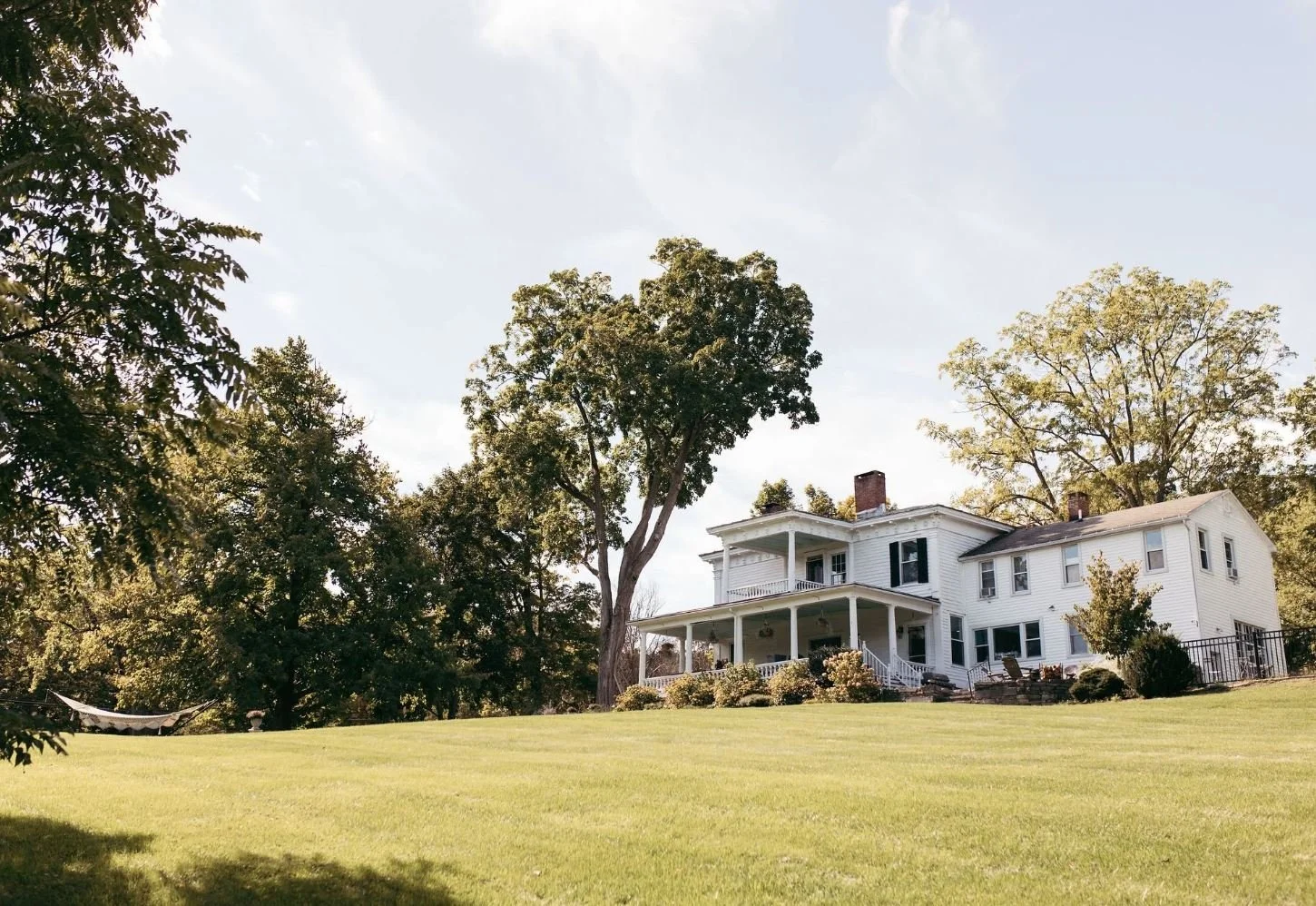A large white two-story house with a wrap-around porch, surrounded by green grass and tall trees under a partly cloudy sky.