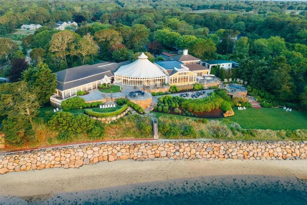 A large house or event venue with a central tent-like structure surrounded by trees and landscaped gardens, located on a coastline with a rocky barrier and sandy beach.