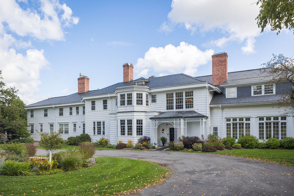 Large white mansion with multiple chimneys, surrounded by a well-maintained garden and a curved driveway.