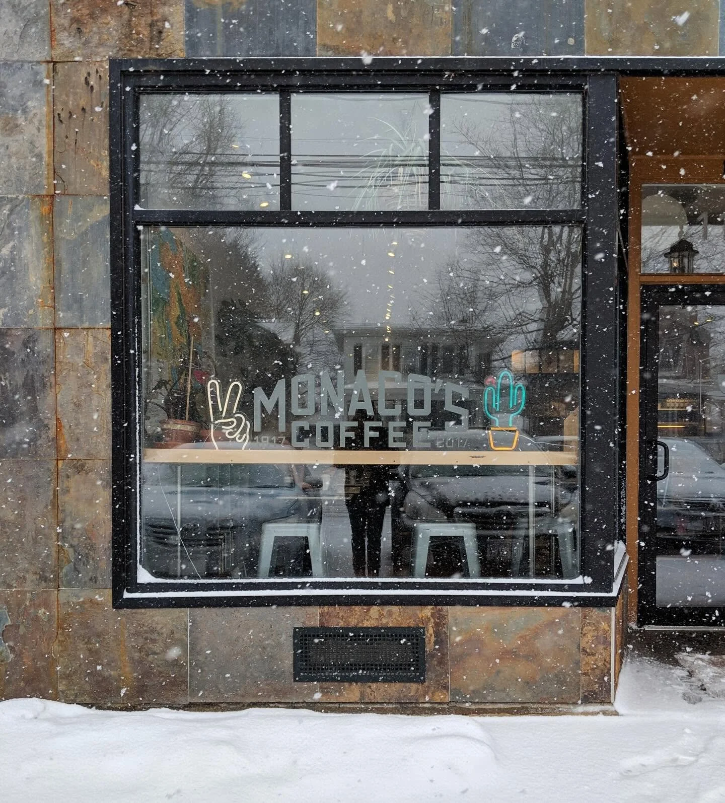 The window of a coffee shop named "Monaco's Coffee" with colorful neon signs of a peace hand gesture and a potted cactus. Snow is falling outside on a snowy sidewalk.