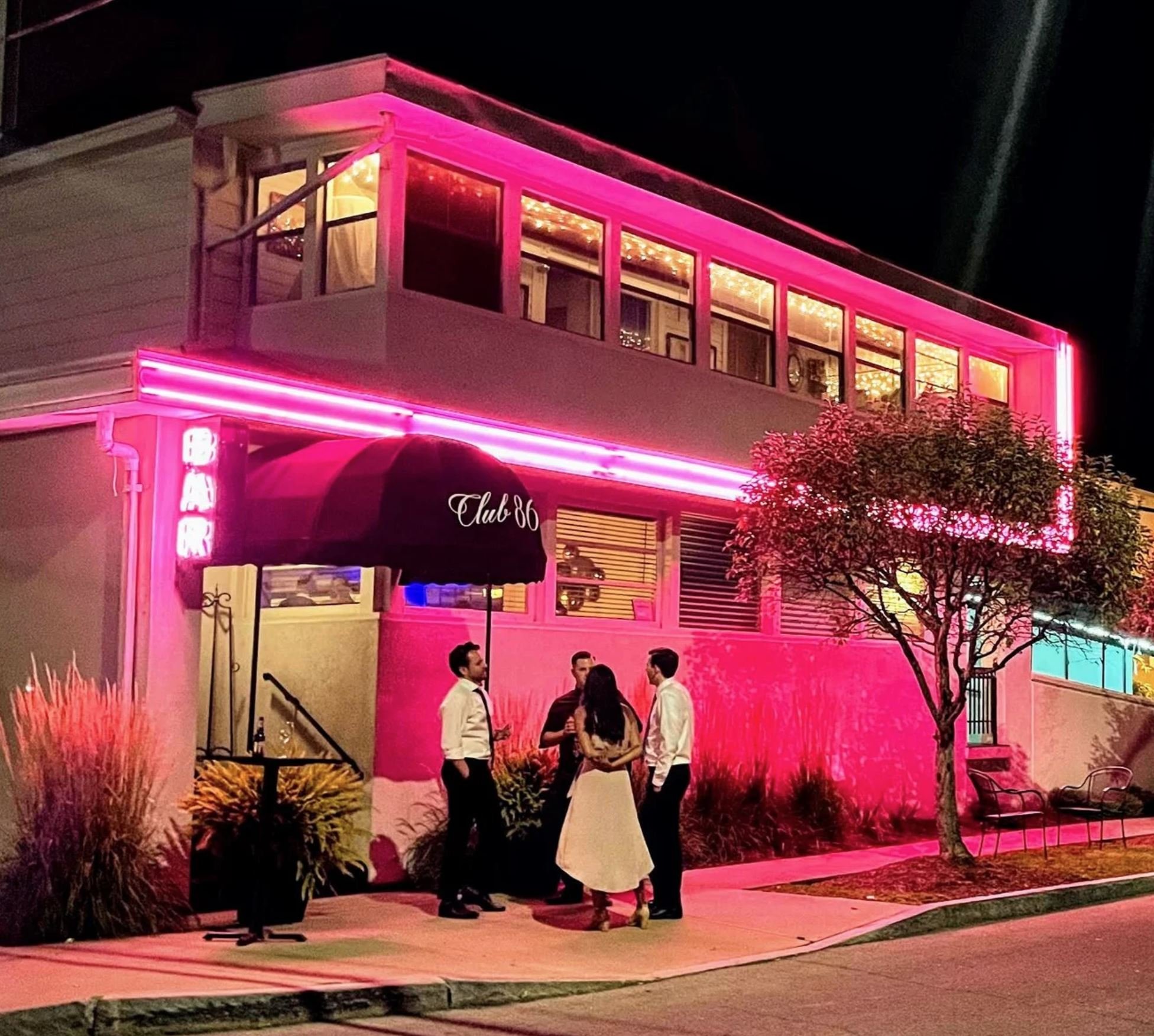 Night scene outside a building with pink neon lights and large glass windows showing warm interior lighting. Several people are gathered, talking in groups. The building has a sign that says 'Club 86' and is decorated with plants and outdoor furniture.
