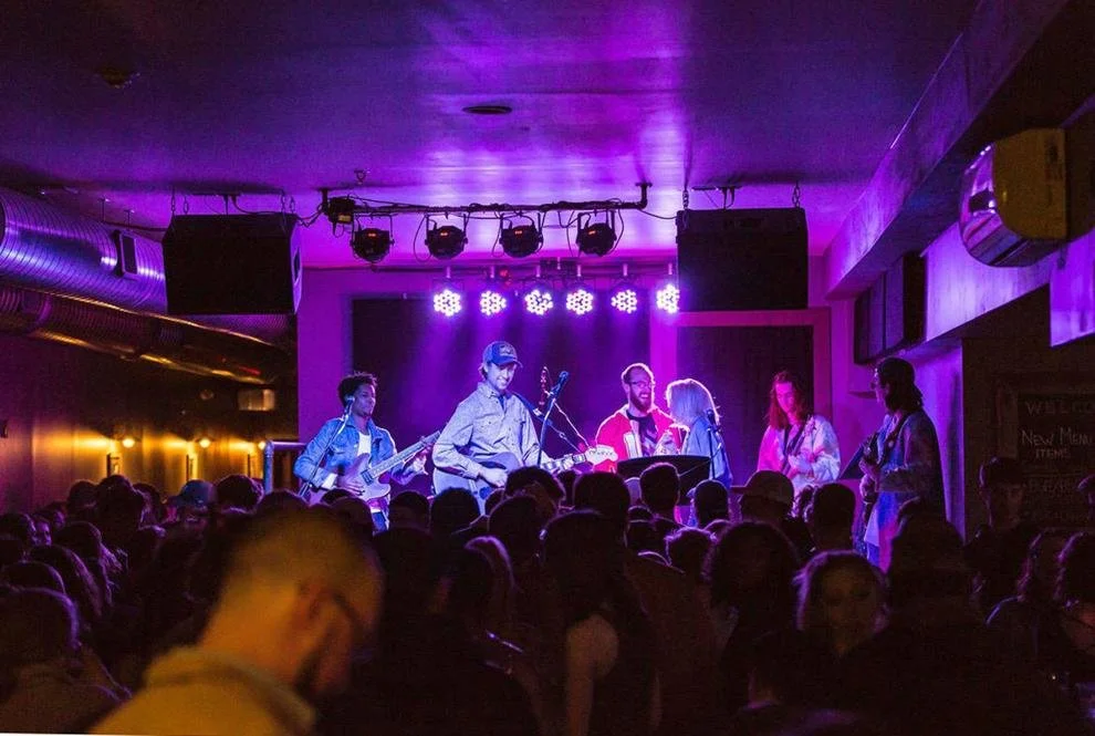 A band performing on stage at a concert with purple lighting, playing guitars and singing, with a crowd of people watching in the foreground.