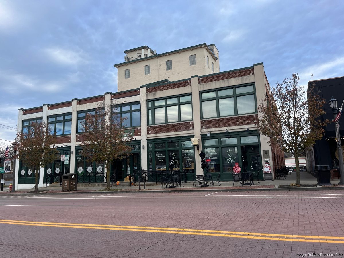 A multi-story building with a ground-level cafe featuring outdoor seating, large green framed windows, trees along the sidewalk, and a street with a brick pavement in front.