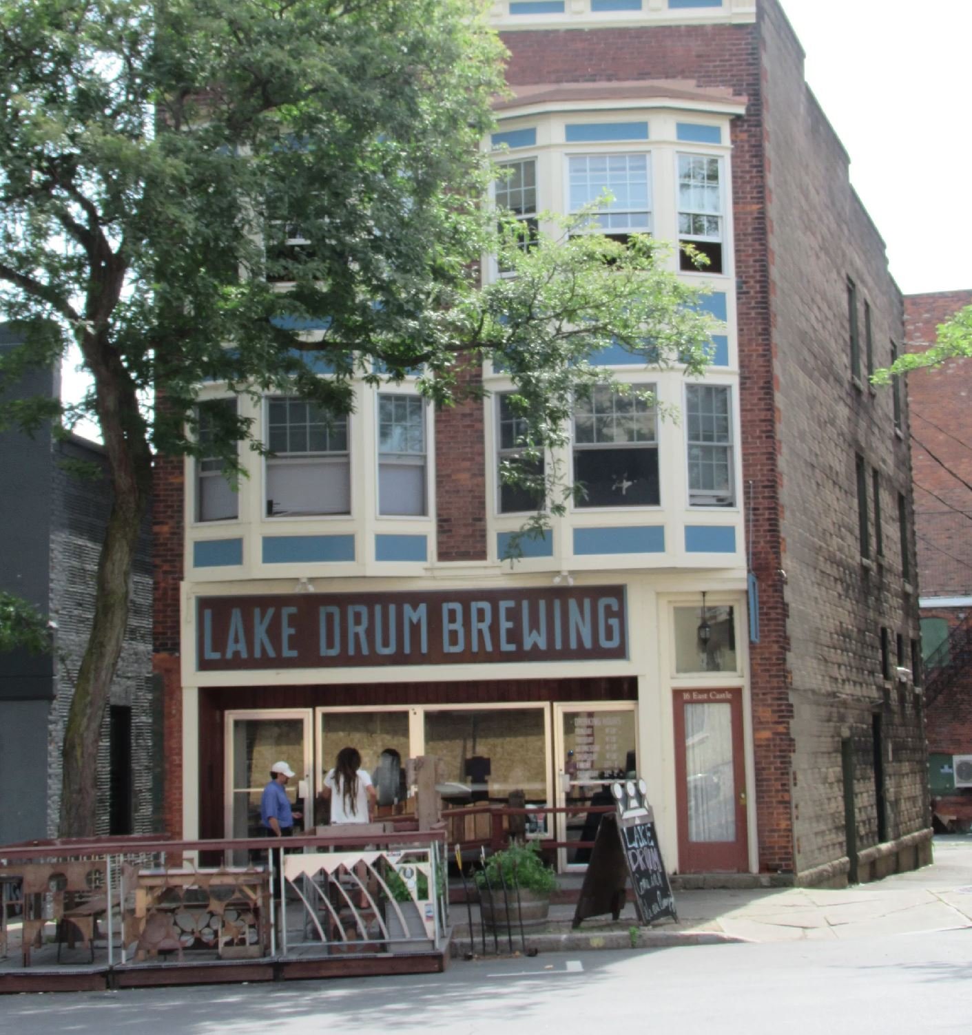A multi-story brick building with a sign that reads "Lake Drum Brewing" on the front. There are large windows on the upper floors and people gathered outside near a wooden railing and a chalkboard menu, with trees partially obscuring the view.