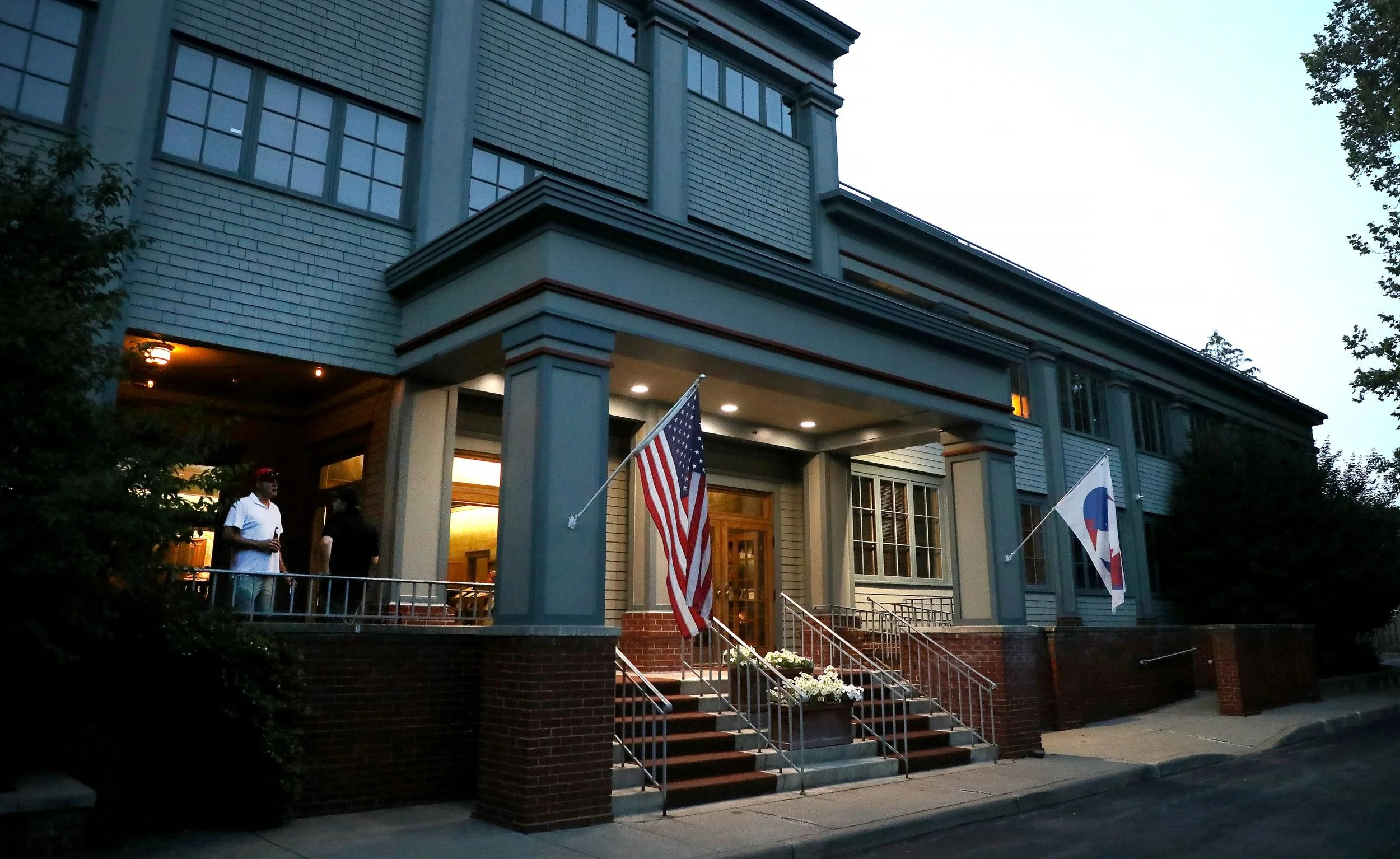 Front view of a large two-story house with a porch, flags, and people standing outside, taken at dusk.