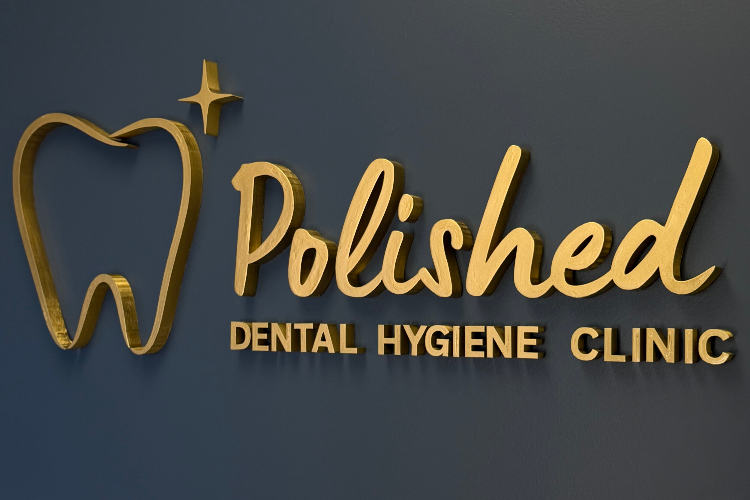 Gold-colored sign with cursive text reading 'Polished' and smaller text below stating 'Dental Hygiene Clinic' on a dark blue wall.