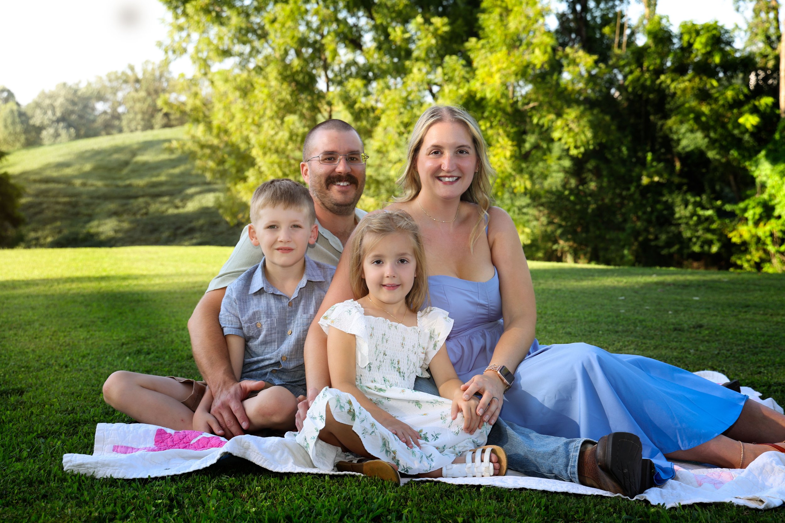 A family of four sitting closely together on a blanket in a park, smiling at the camera with green trees and grass in the background.