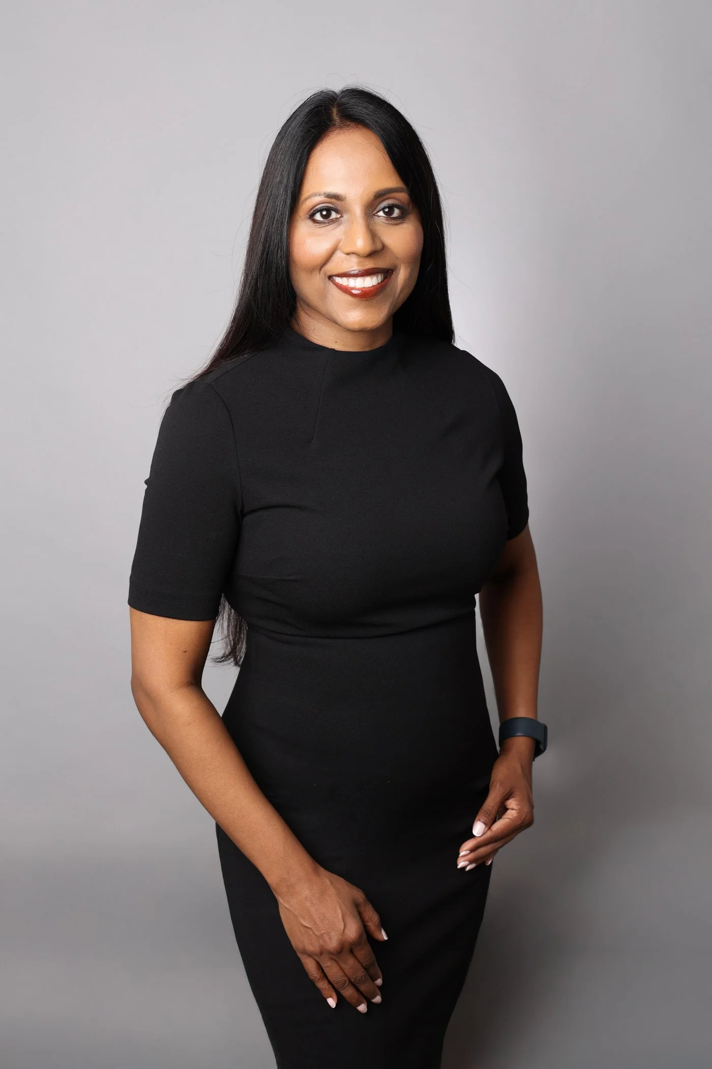 A woman with long black hair smiling, wearing a black dress and a fitness tracker on her left wrist, standing against a plain gray background.