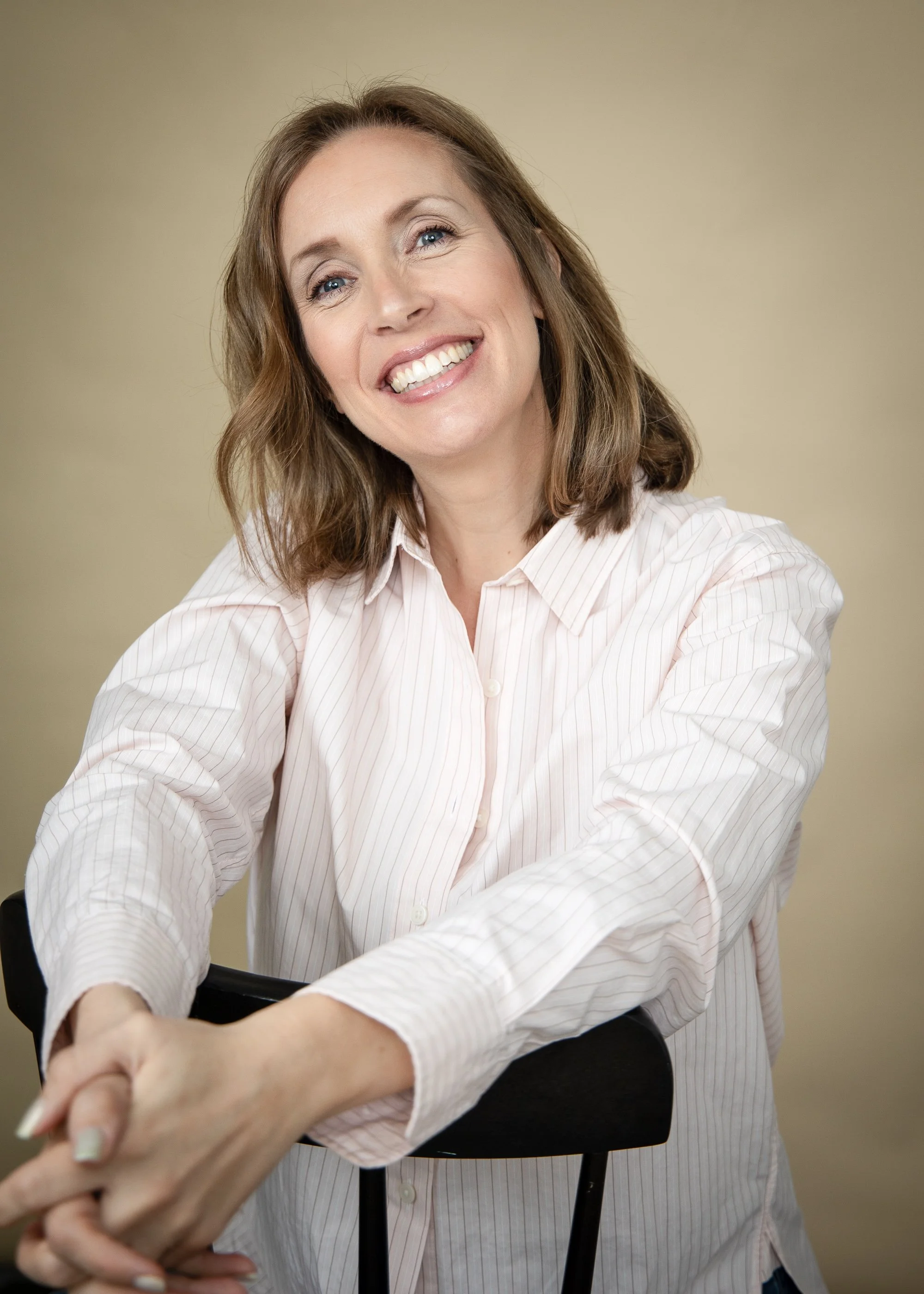 Woman smiling while holding hands with someone, sitting on a black chair, in front of a beige wall.
