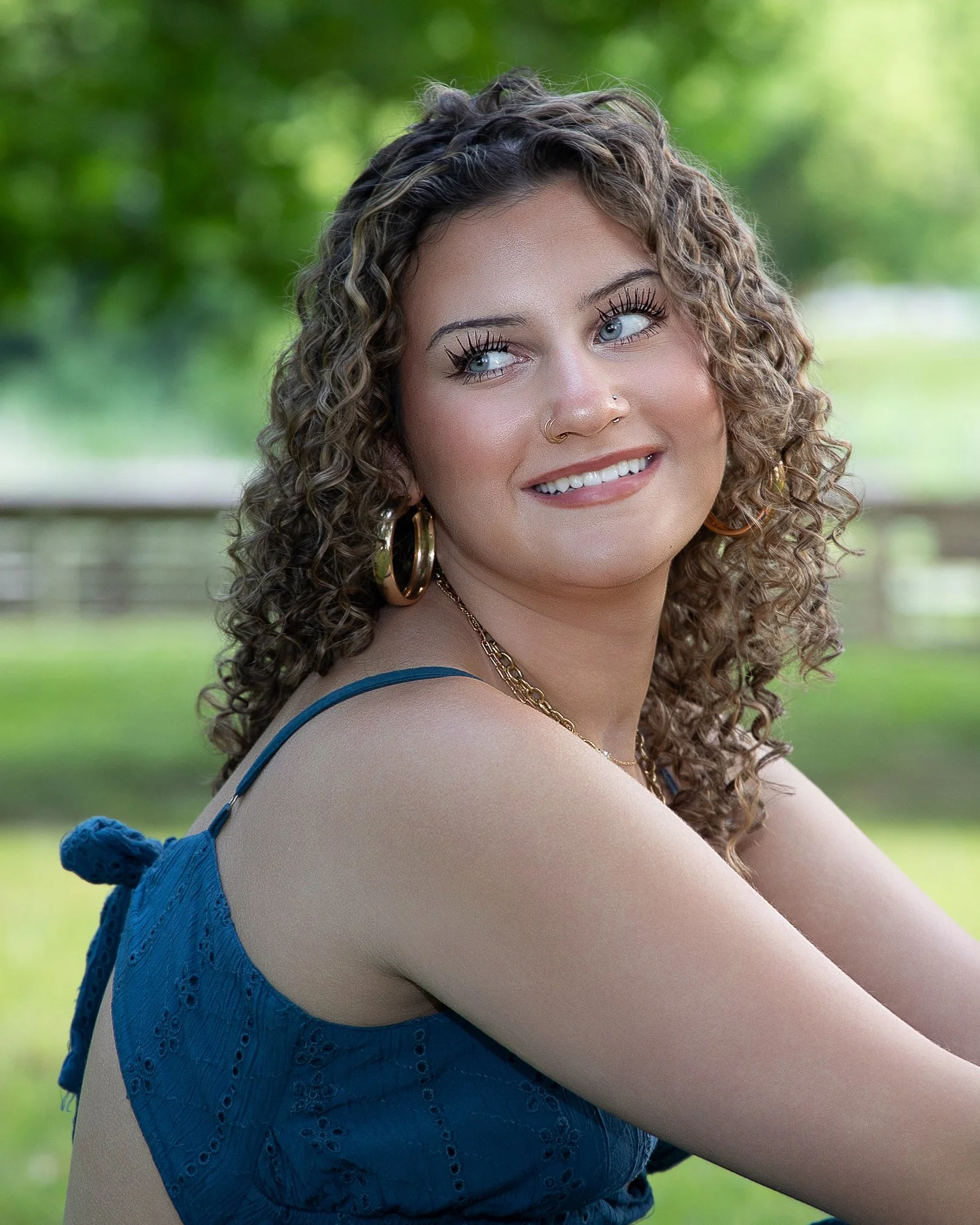 Close-up of a young woman with curly hair, blue eyes, and jewelry, smiling outdoors with a blurred green background.