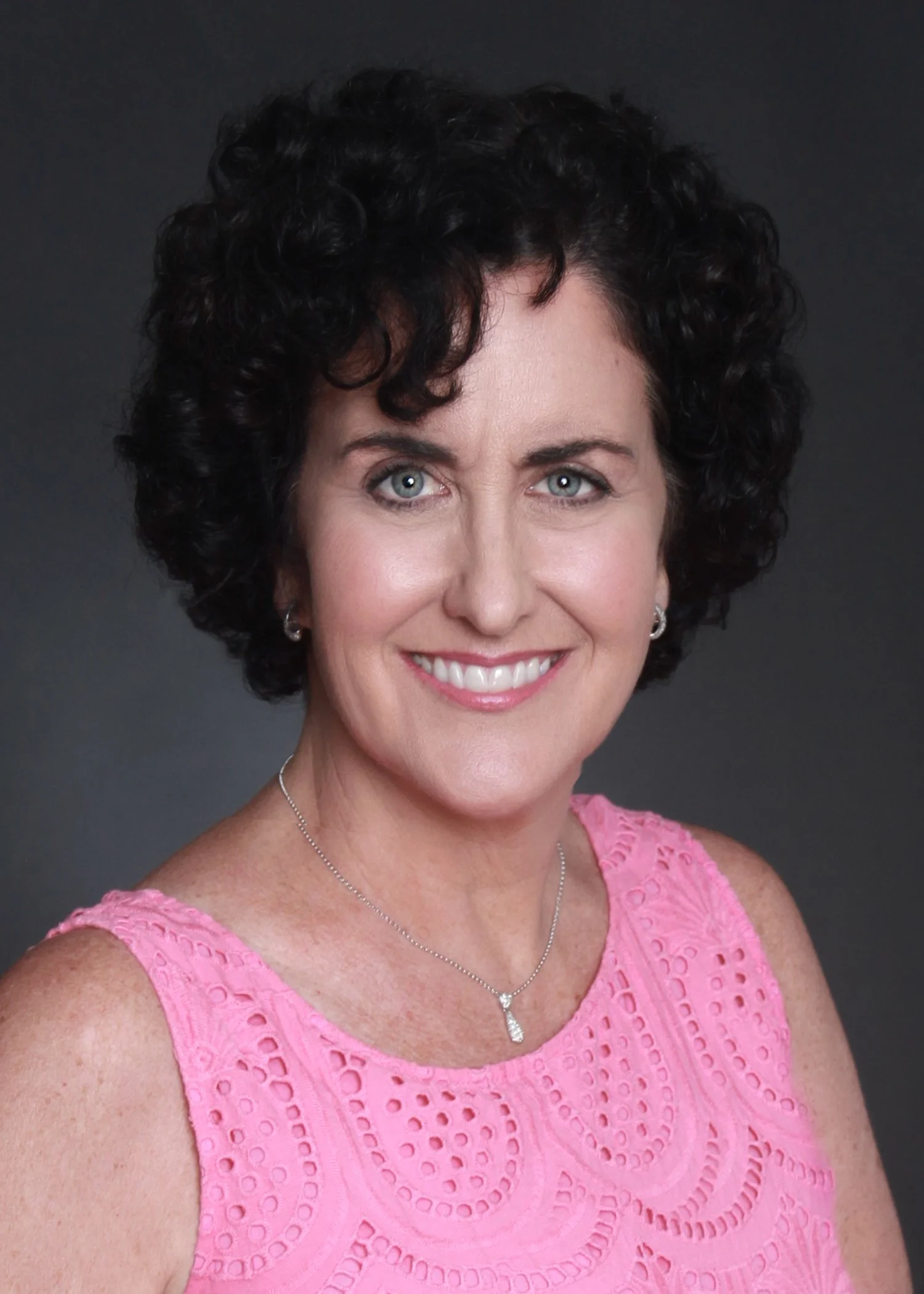 A smiling woman with short, curly dark hair, wearing a pink sleeveless dress with cutout patterns, a silver necklace with a pendant, and matching earrings, against a dark background.