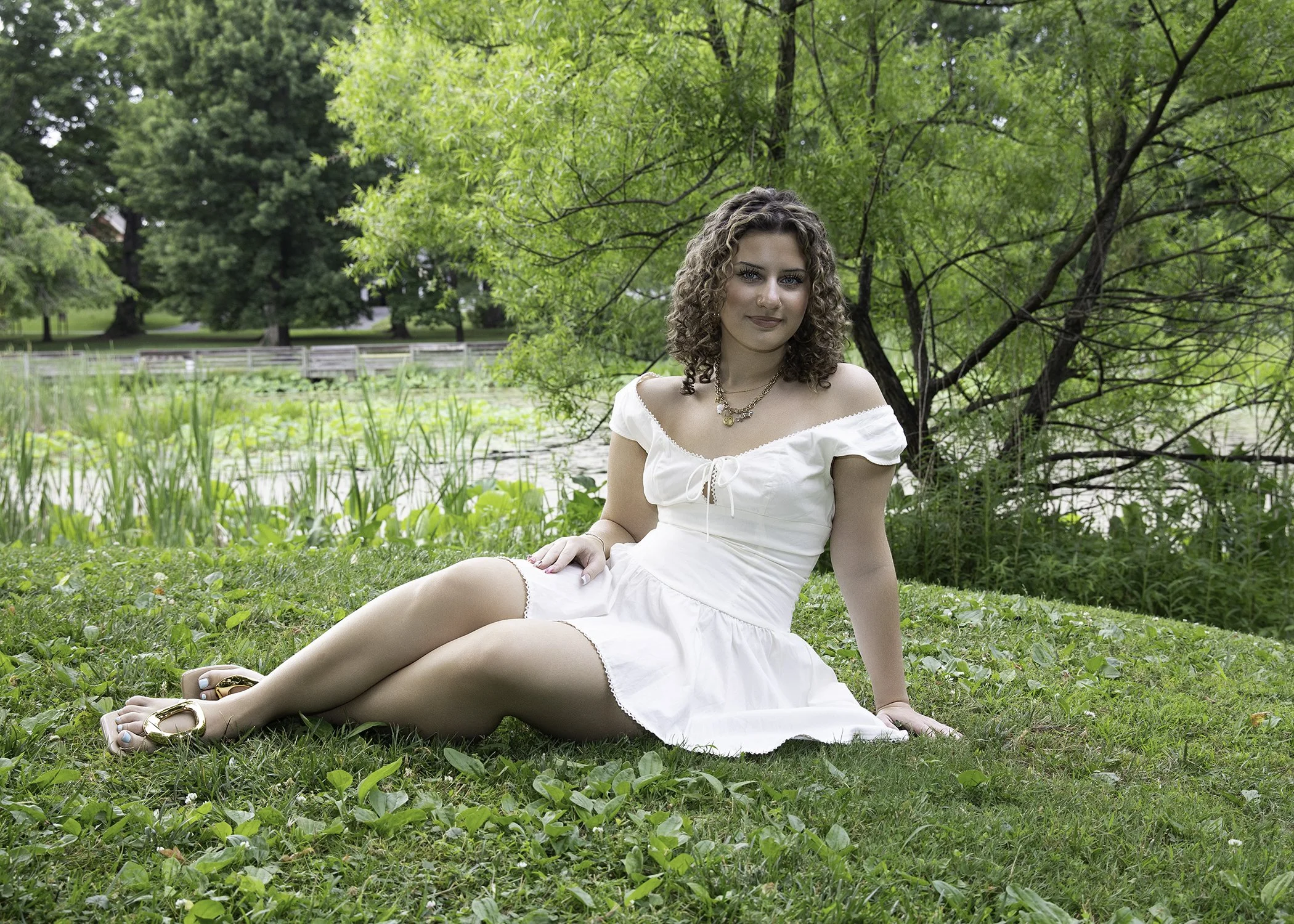 A woman with curly hair sitting on grass near a pond, surrounded by green trees, wearing a white dress, gold jewelry, and sandals in a natural setting.