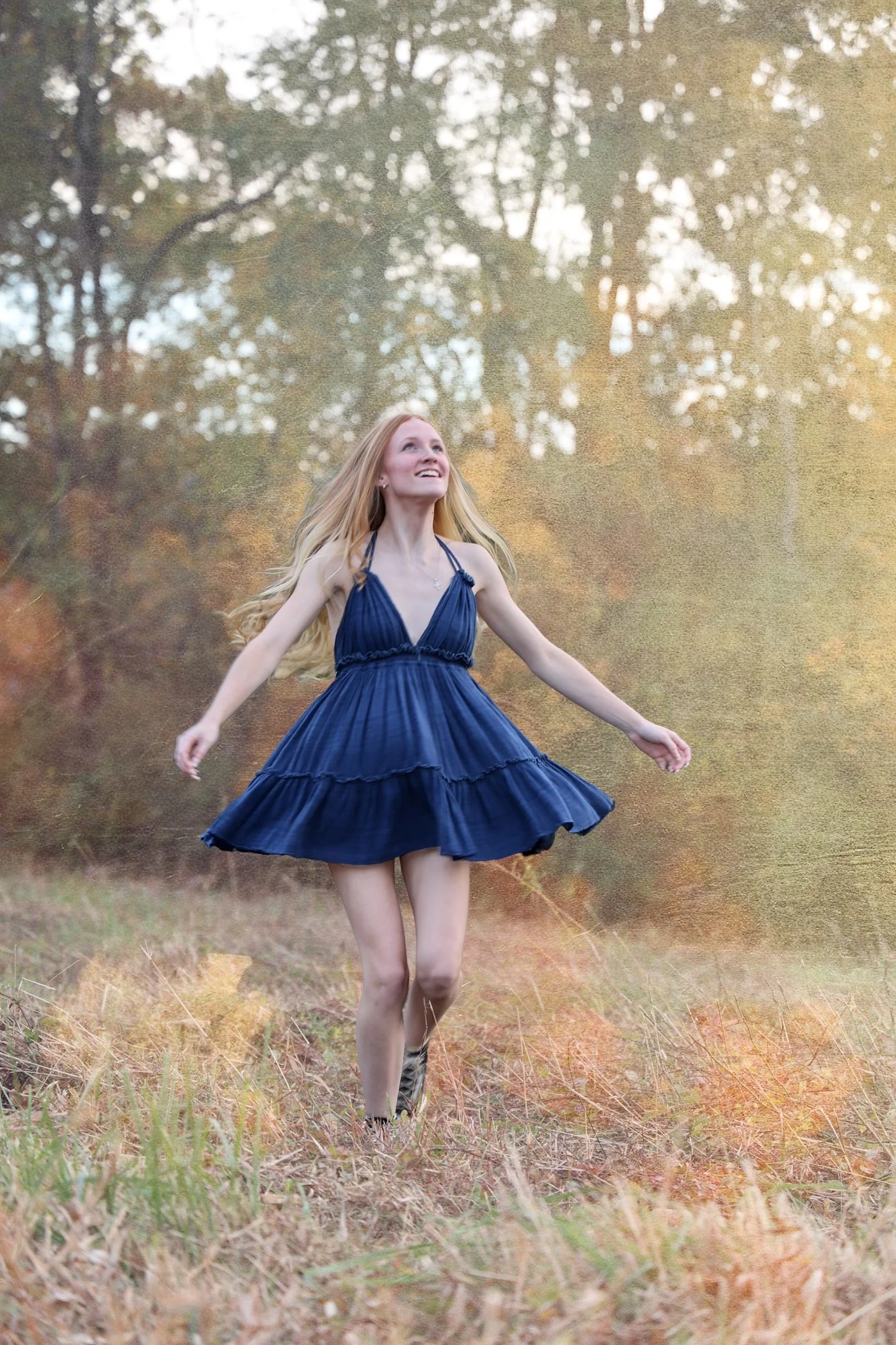 A young woman with long blonde hair wearing a sleeveless navy blue dress dancing outdoors in a grassy field with trees in the background.