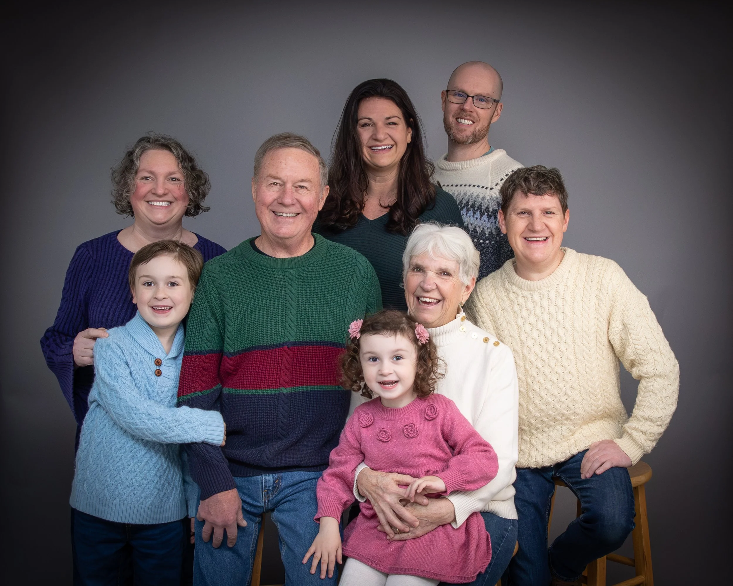 A multi-generational family portrait with seven smiling people, including children, middle-aged, and elderly members, casually dressed, standing against a gray backdrop.